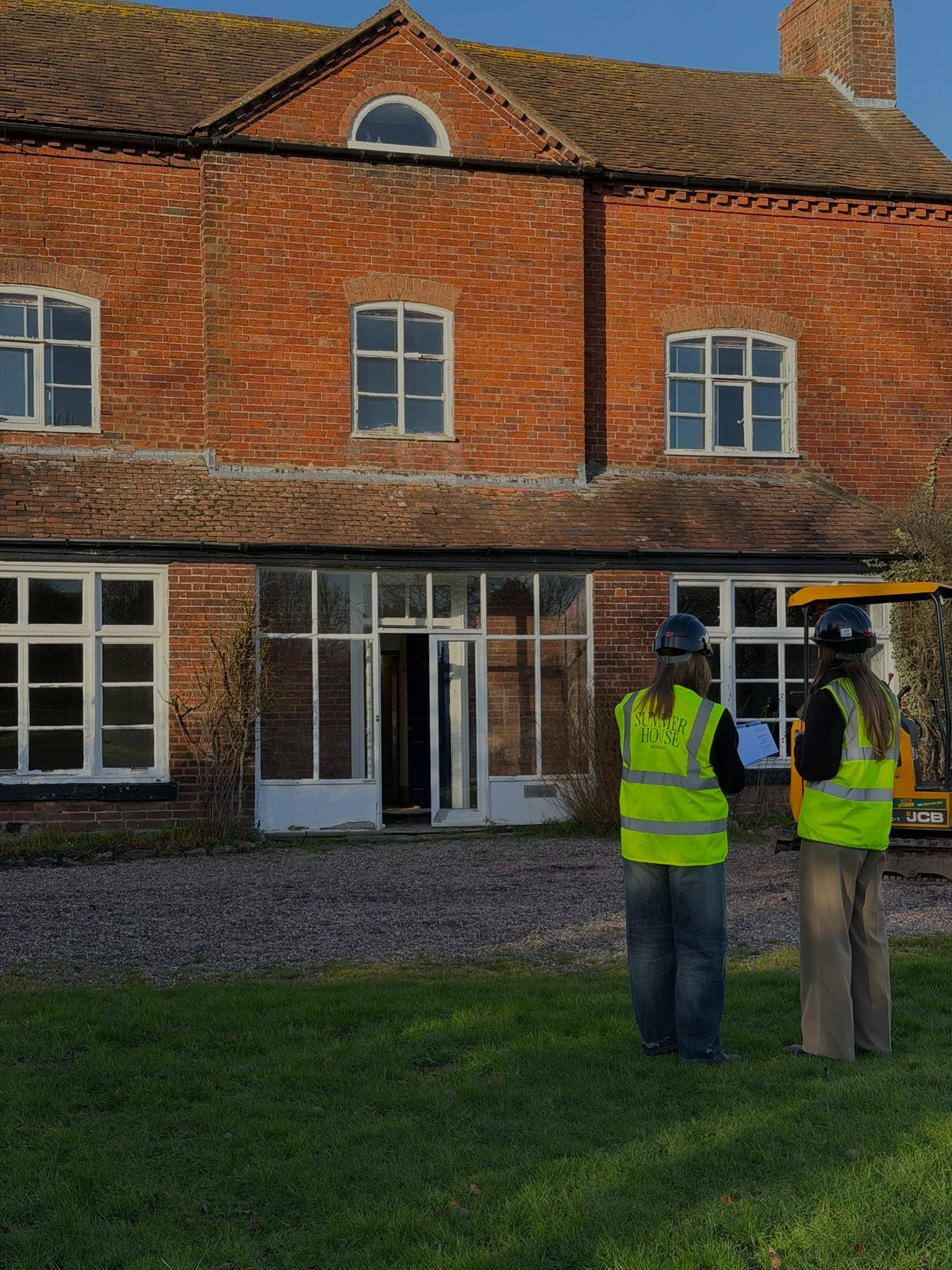 Two women wearing yellow safety vests and helmets standing outdoors in front of a brick house, discussing or inspecting something. The house has multiple large windows and a wooden door.