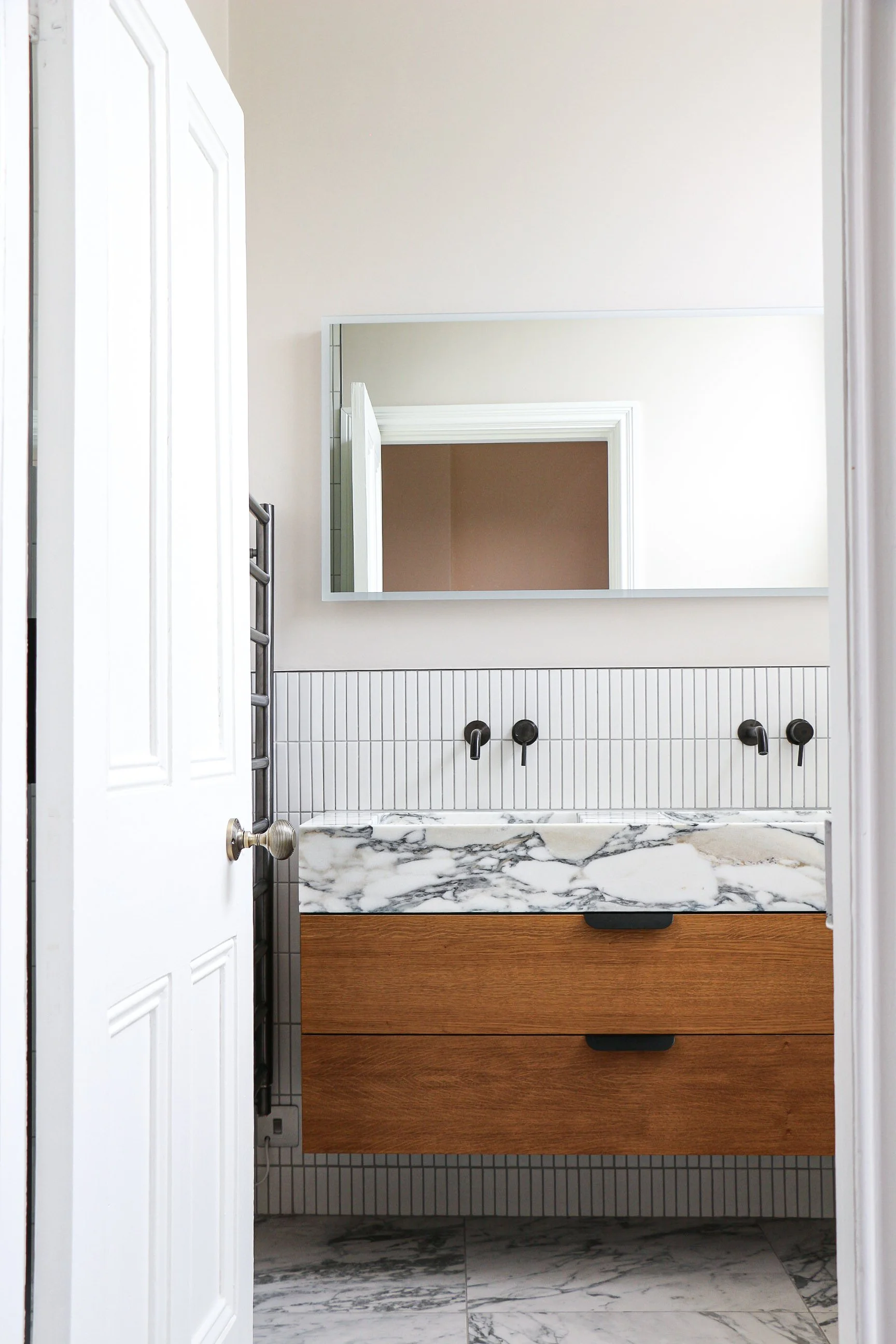 Bathroom mirror above a marble sink with wooden cabinet drawers, white tiled wall with black fixtures, and a window reflected in the mirror.