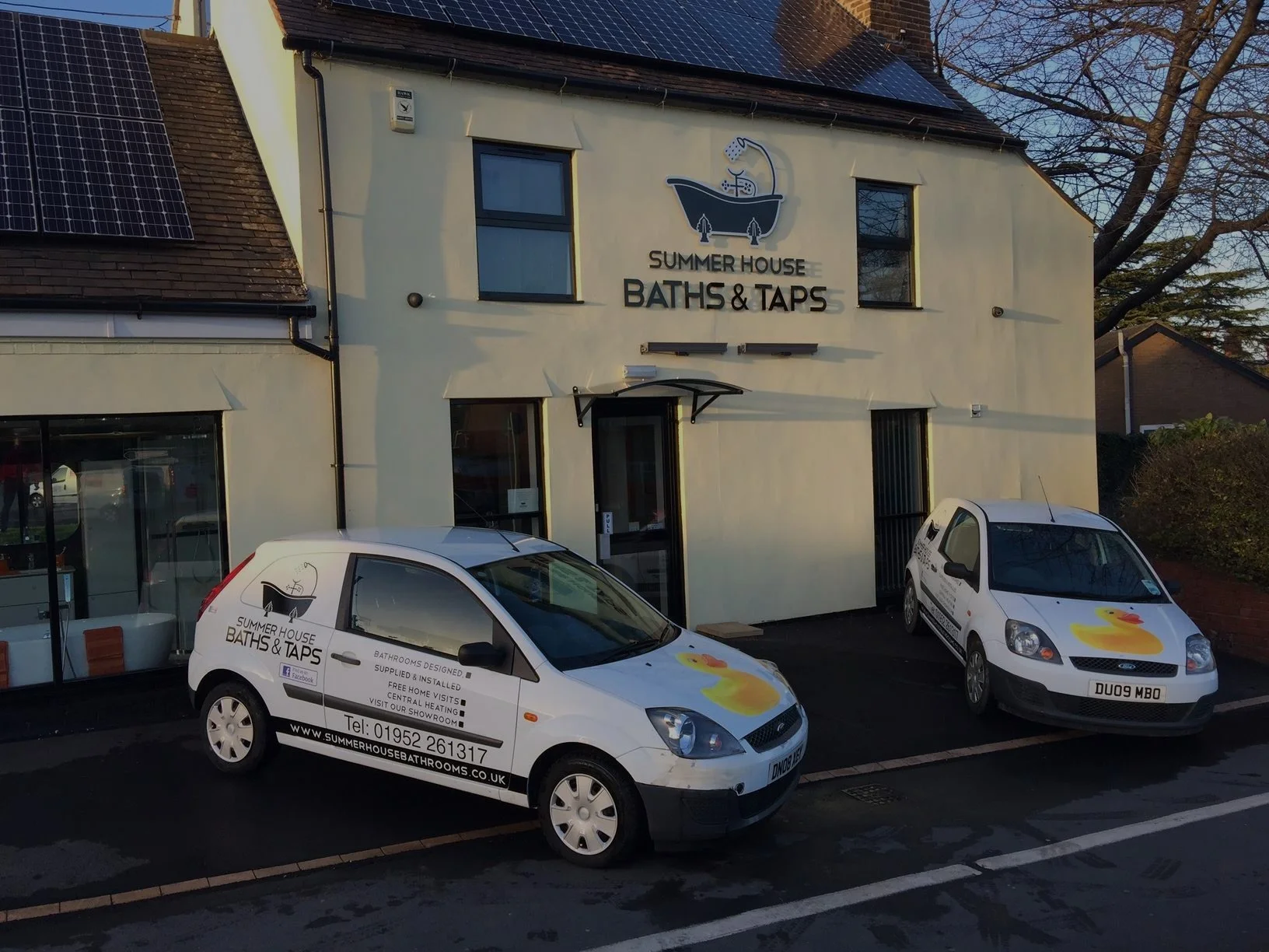Front of a building with signage for Summer House Baths & Taps, cars parked in front, including one with advertising for the business.
