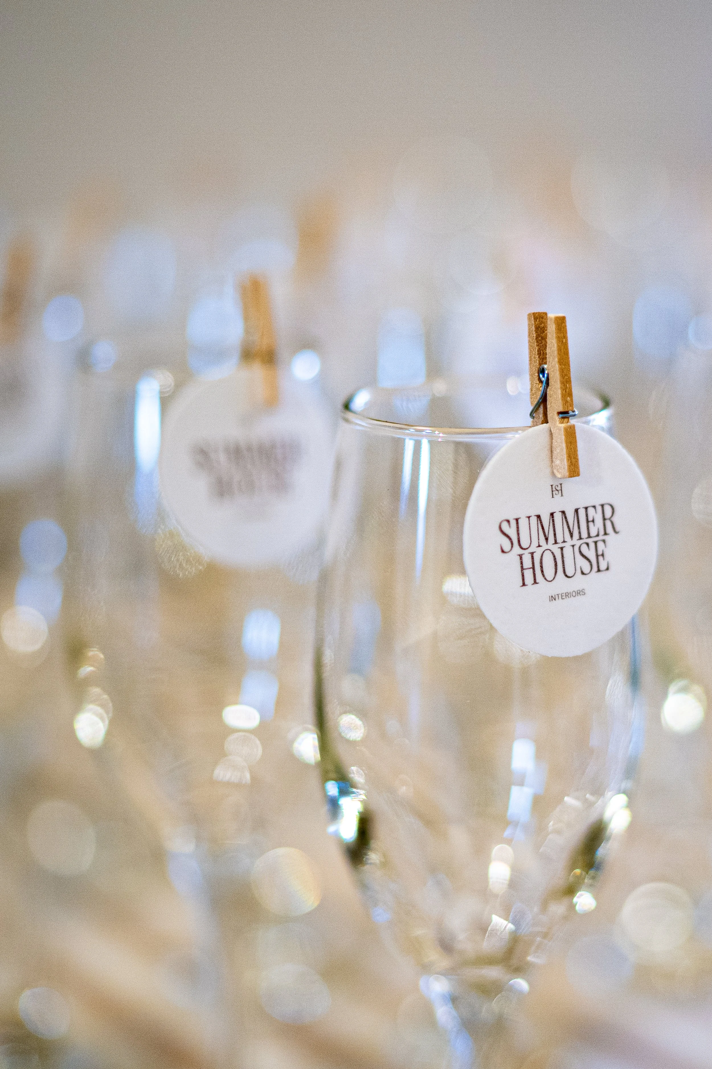 A close-up photograph of a small glass container with a white tag that reads 'Summer House Interiors' attached to a wooden clothespin, with a blurred background of lights.
