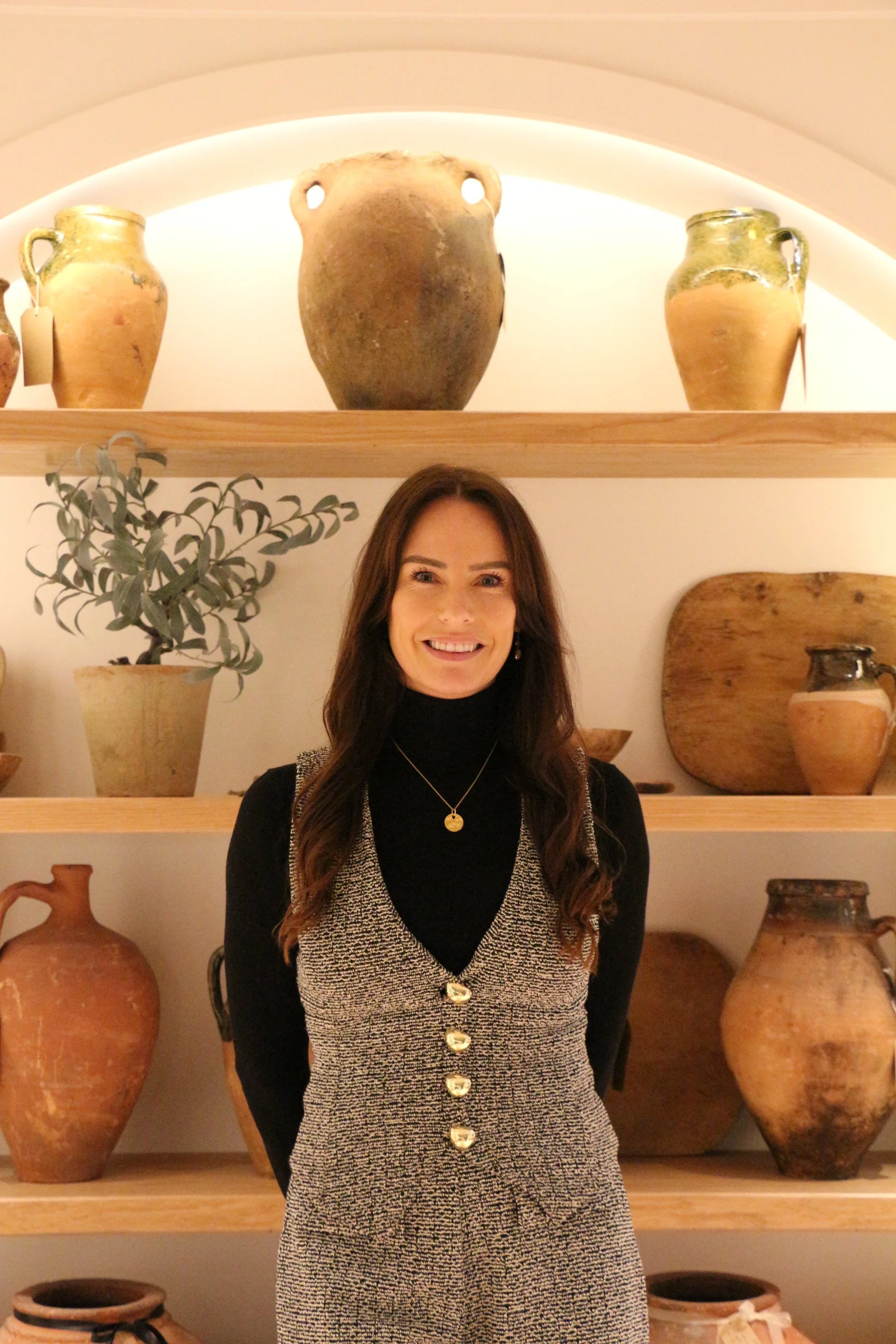 A smiling woman with long brown hair standing in front of a shelf with large ancient clay pots and a potted plant, in a well-lit room with arched ceiling.