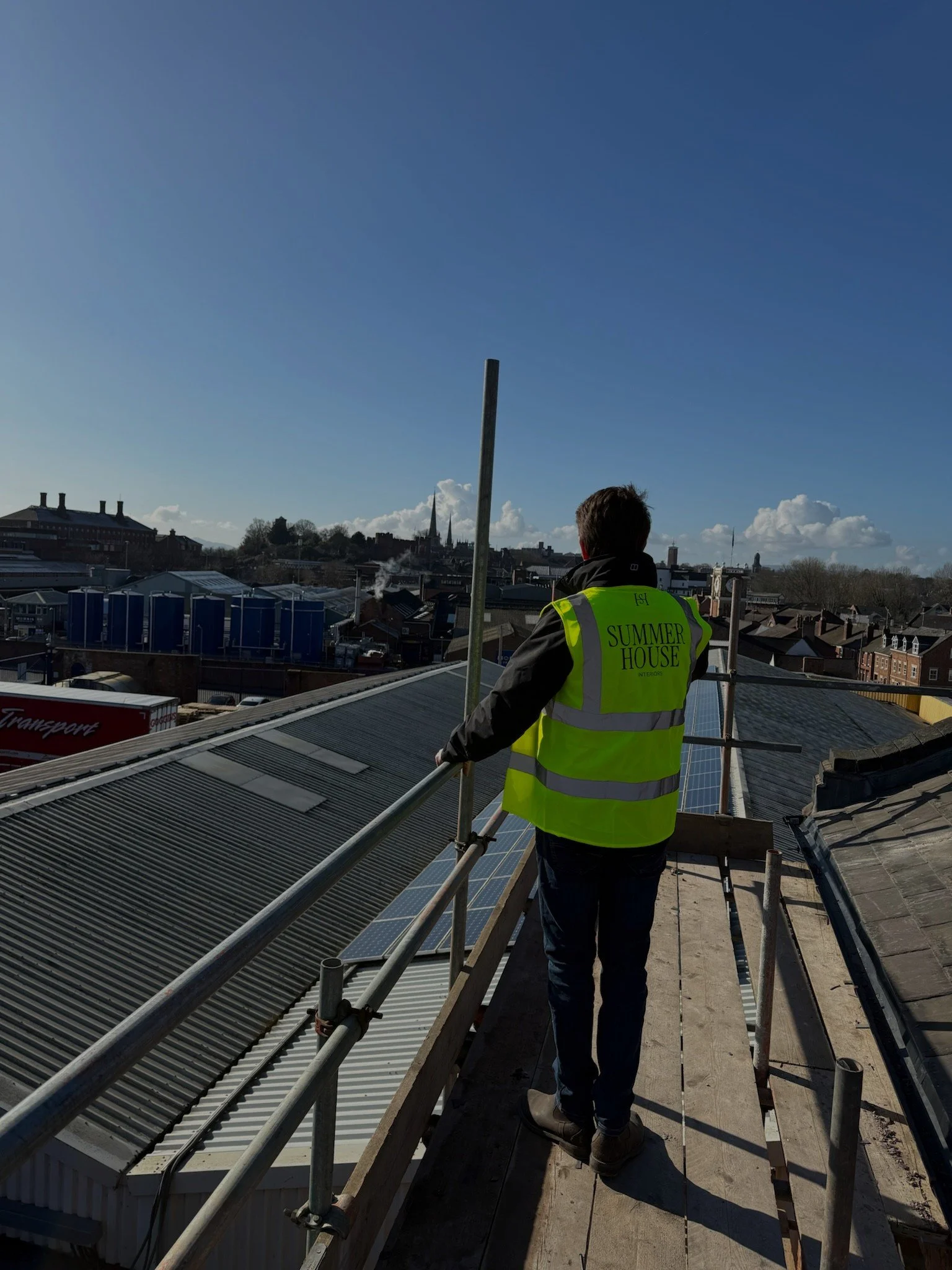 Person in a yellow high-visibility safety vest standing on a rooftop in daytime, overlooking buildings with solar panels and a city skyline in the background.