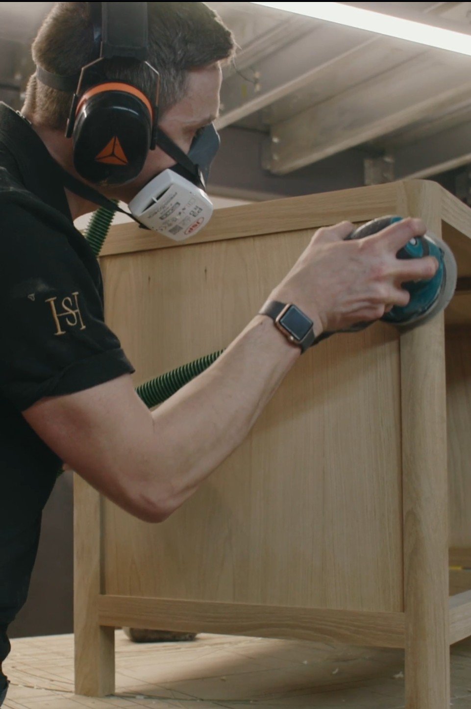 A man wearing a respirator mask and eye protection is sanding a wooden piece of furniture with a power sander.