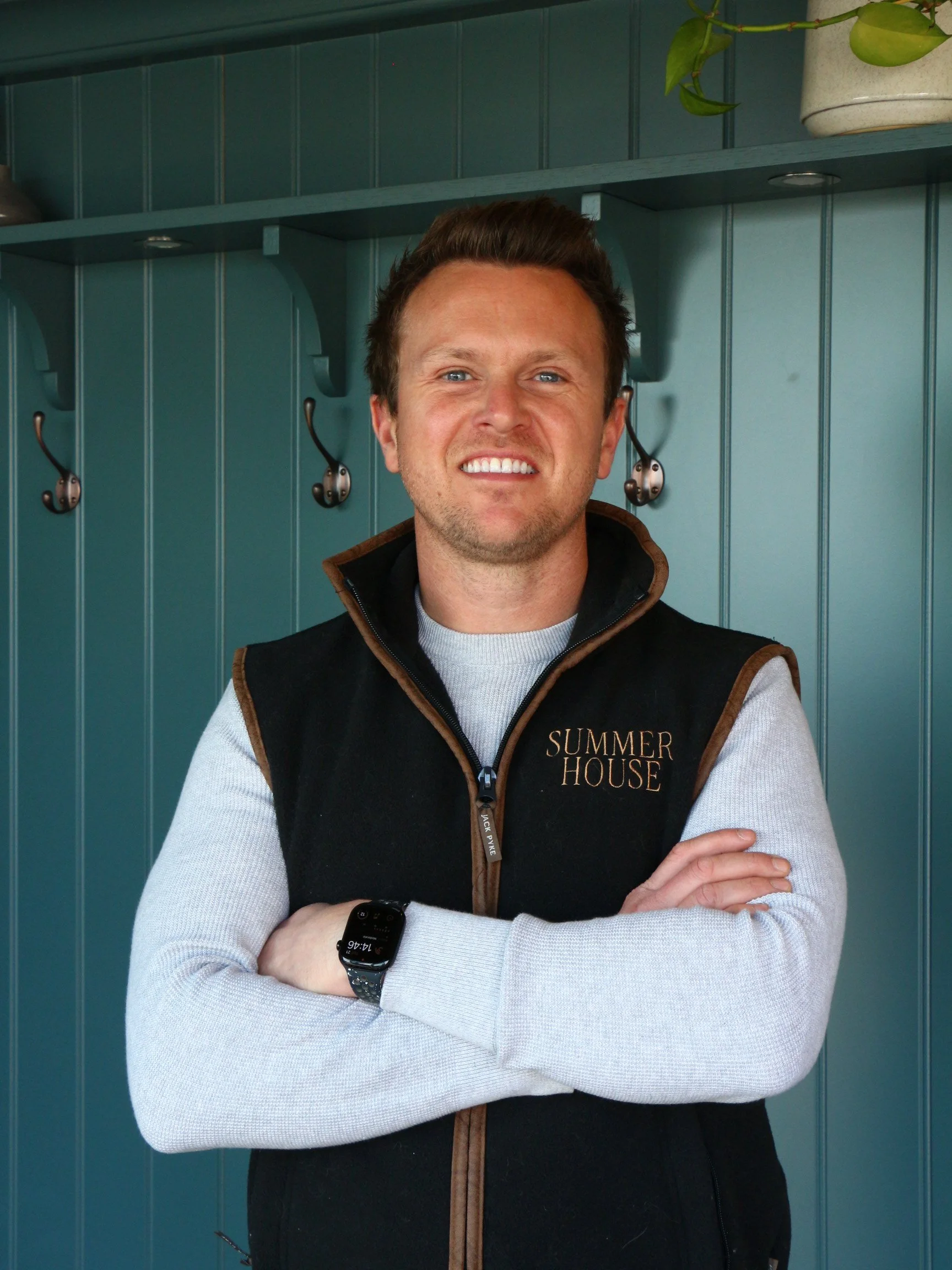 A smiling man with blue eyes, wearing a gray turtleneck and a black vest with brown trim that has the words 'Summer House' embroidered on it, standing in front of a teal wooden wall with hooks and a potted plant.