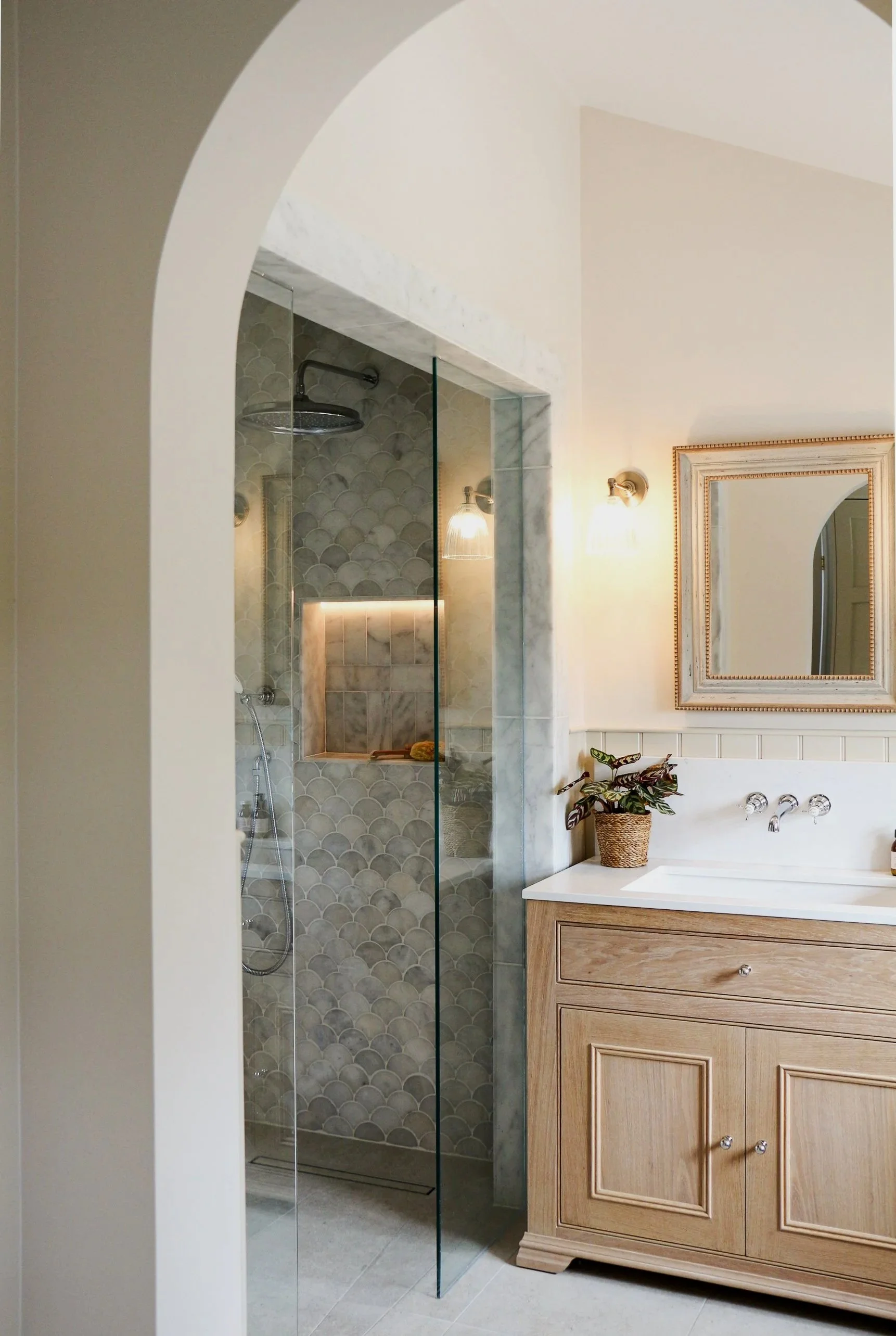 Bathroom with a walk-in shower featuring scalloped tile walls, a rainfall showerhead, a handheld shower, a mirror with a gold frame, a vanity with a wooden cabinet, a white countertop, a potted plant, and wall-mounted lights.