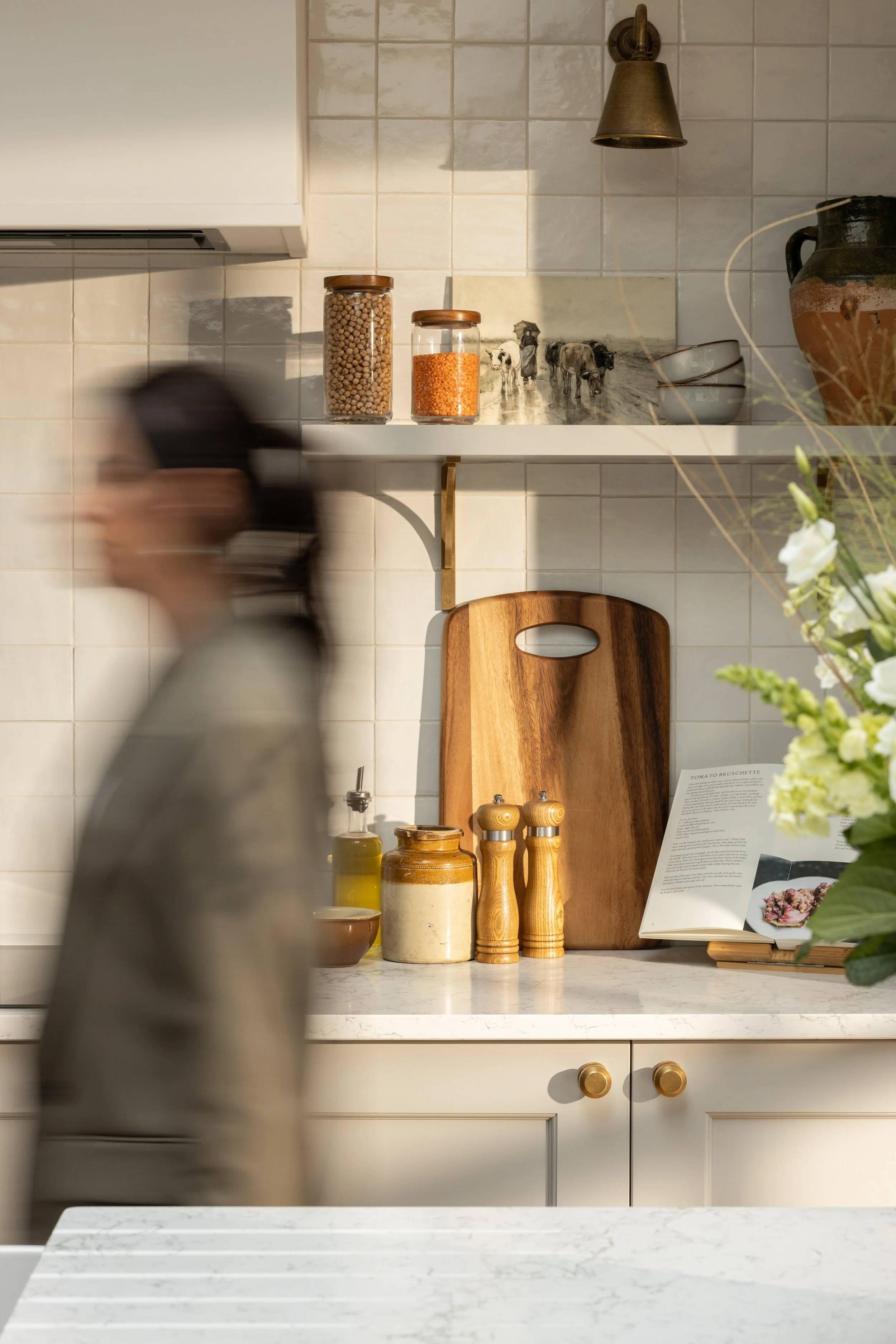 A kitchen counter with jars of spices, a cutting board, a cookbook, and a flower vase, with a person walking past in a blur.