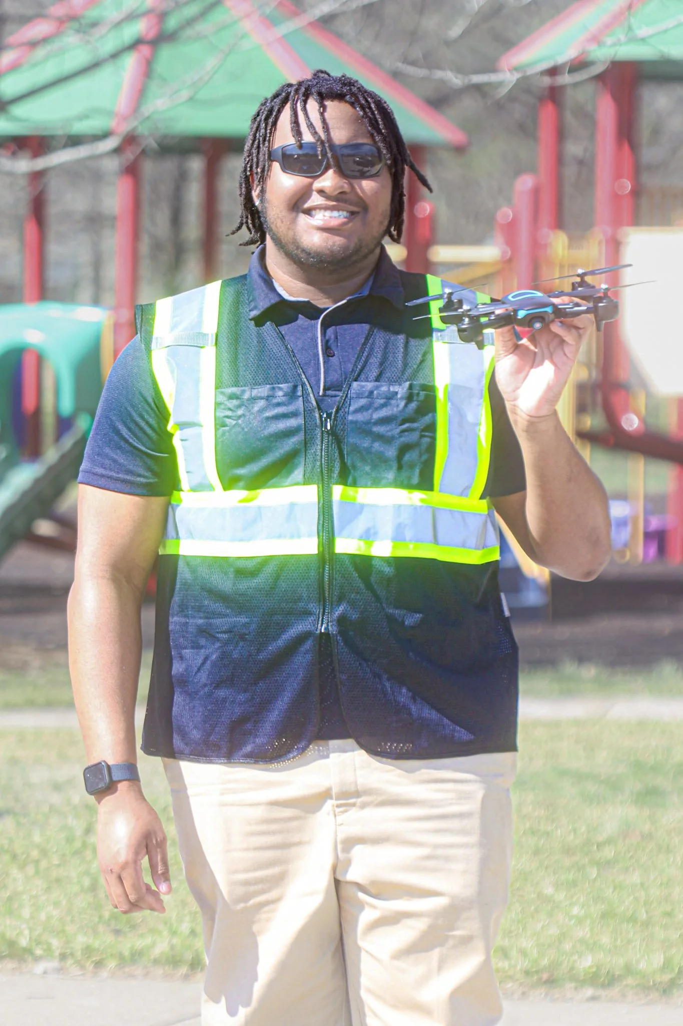 Man wearing sunglasses and a safety vest holding a drone outdoors, with a playground in the background.