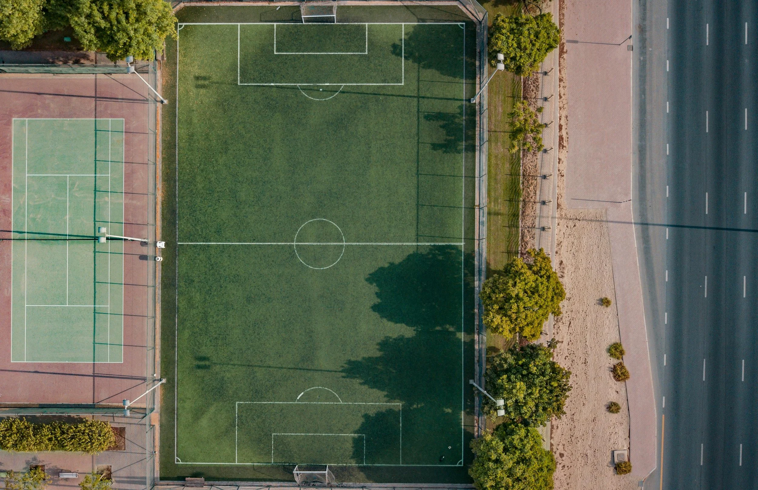 Aerial view of sports courts and a soccer field. The soccer field has a grass surface with white markings and two goals, surrounded by a fence. Adjacent to it are tennis courts with green surfaces, also enclosed by a fence, and lit by tall outdoor lights. To the right of the fields is a sidewalk with trees and nearby a multi-lane road.