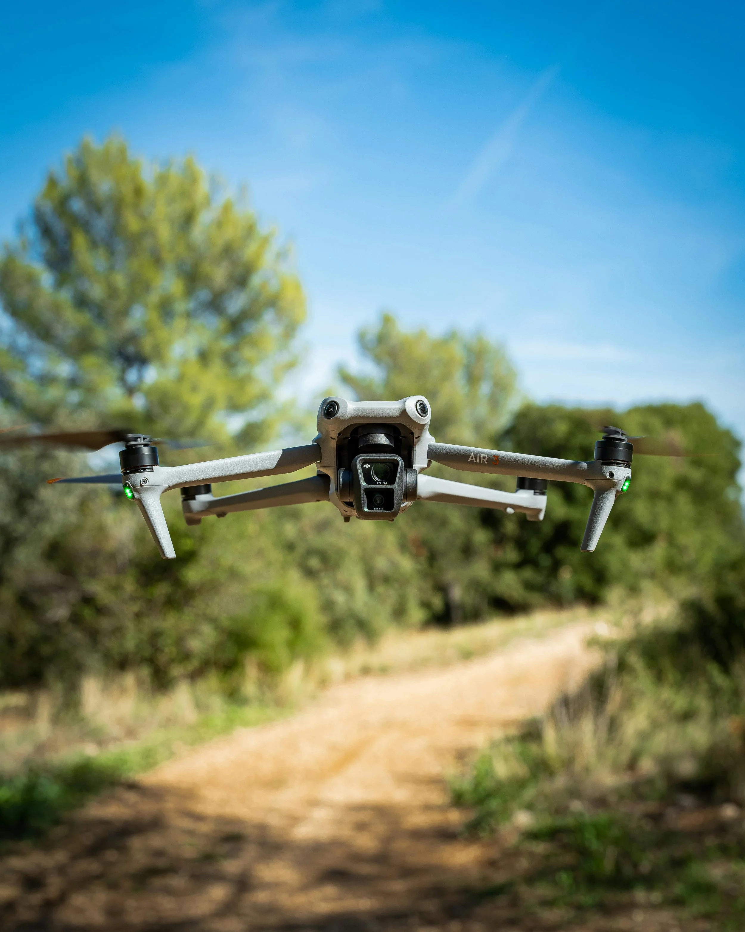 A drone flying in the sky above a dirt trail surrounded by green trees and a blue sky.