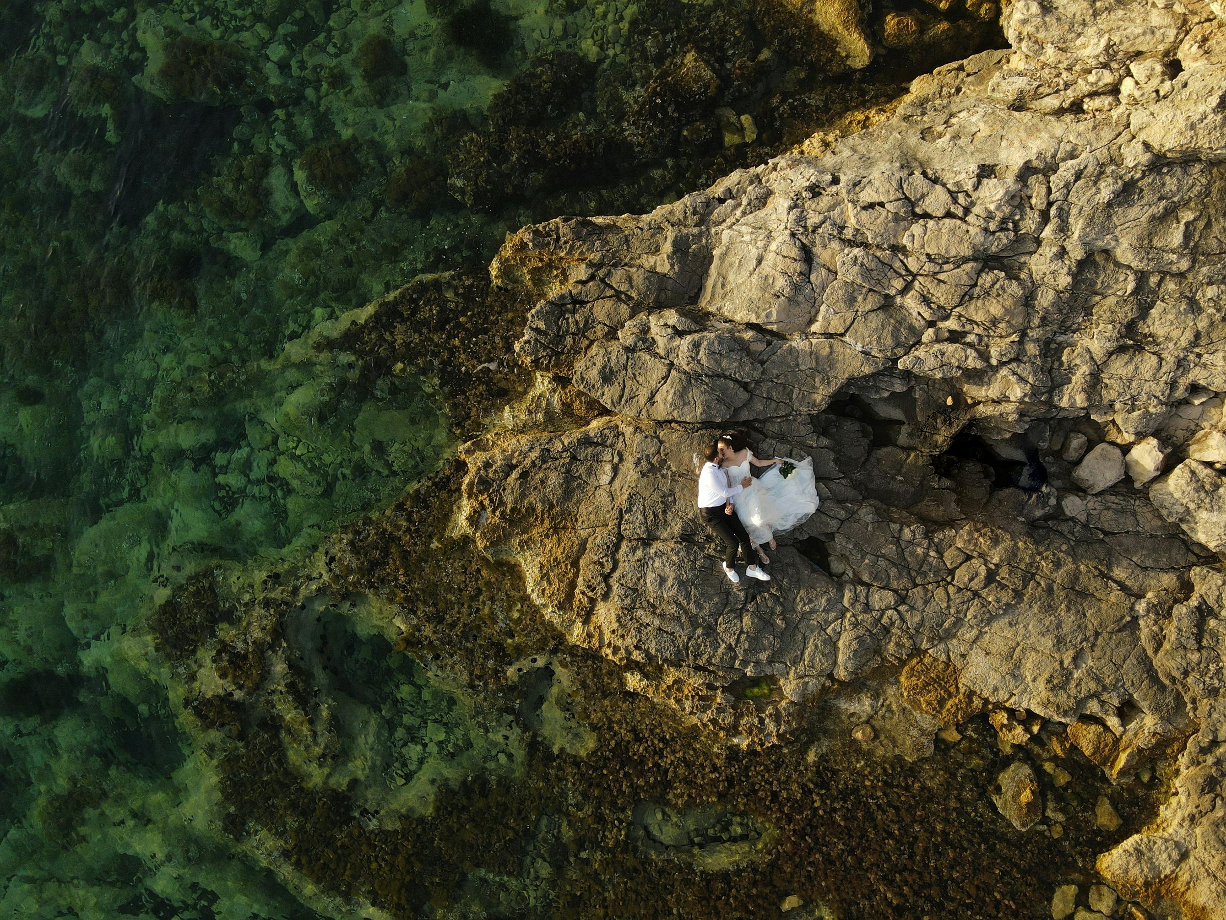 A couple dressed in wedding attire lying on a large rocky formation near a clear green body of water.