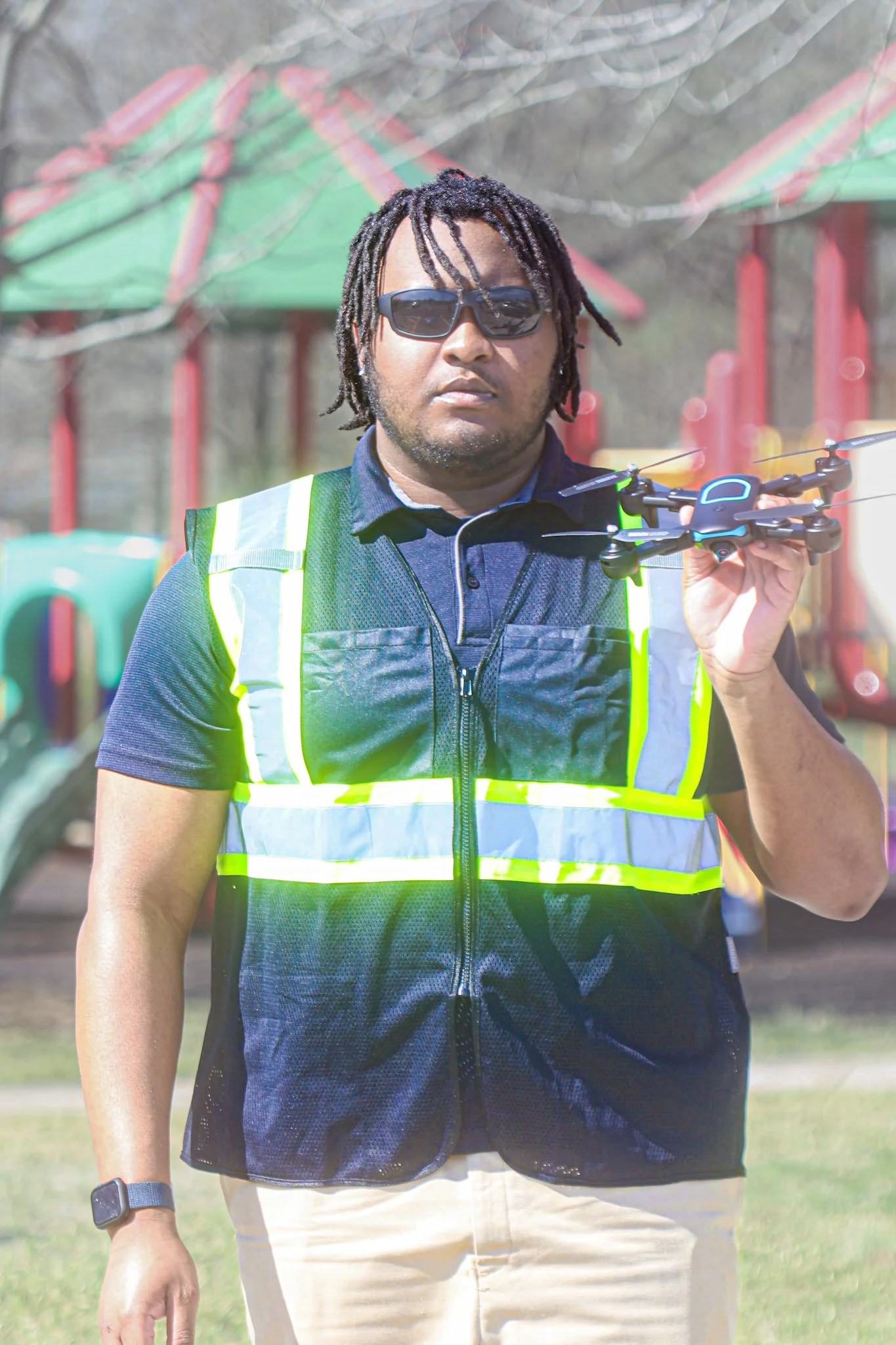 Man with dreadlocks, sunglasses, wearing a reflective vest, holding a drone in an outdoor park with playground equipment.