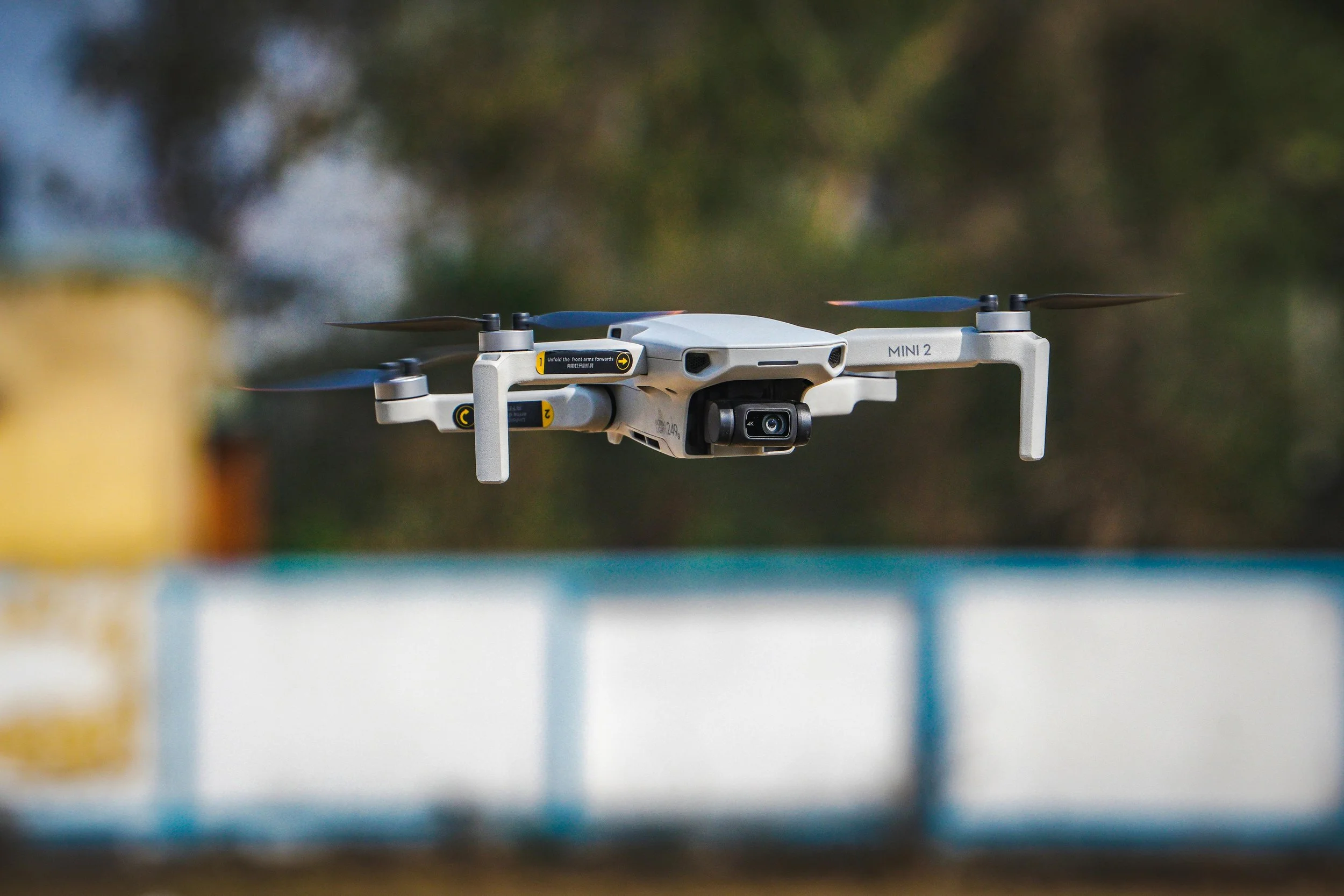 A DJI Mini 2 drone flying outdoors at a low altitude with a blurred background of trees and a sky.