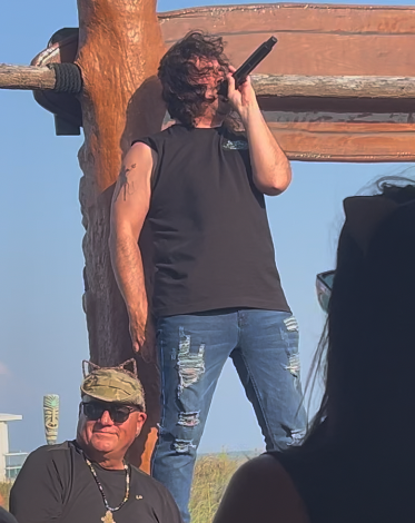 Lead singer in a black sleeveless shirt, holding a microphone, standing on the railing with a clear blue sky in the background. Another person with a hat and sunglasses is in the foreground.