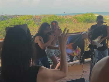 A group of people enjoying a live outdoor music performance near the coast, with some taking photos and others playing instruments.