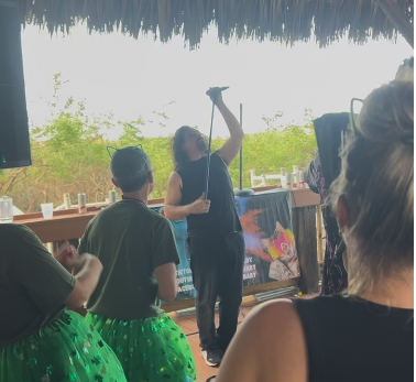 The band performing with people watching at an outdoor event with a thatched roof and trees in the background.