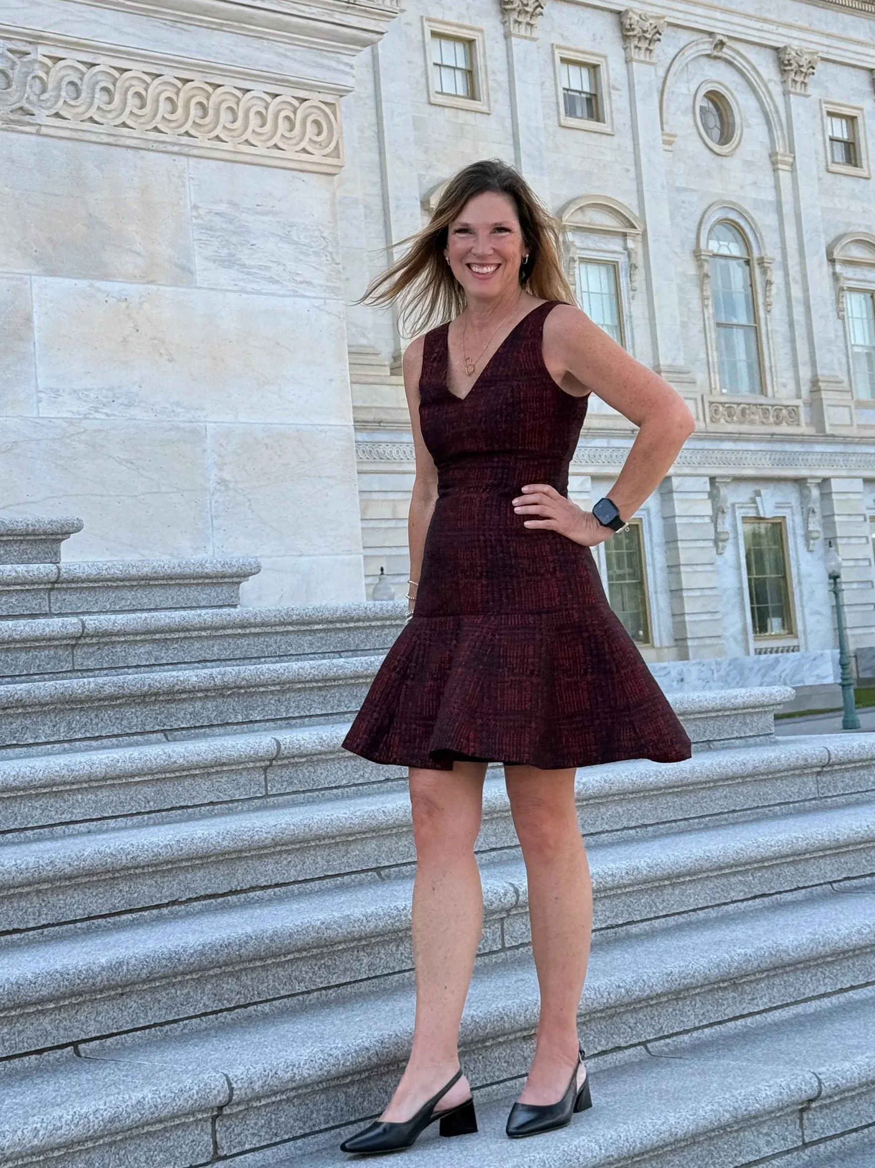 A woman in a sleeveless maroon dress with a flared skirt, black high heels, wearing a watch and a necklace, standing on stone steps outside a historic building with large windows and decorative architectural details, smiling at the camera.