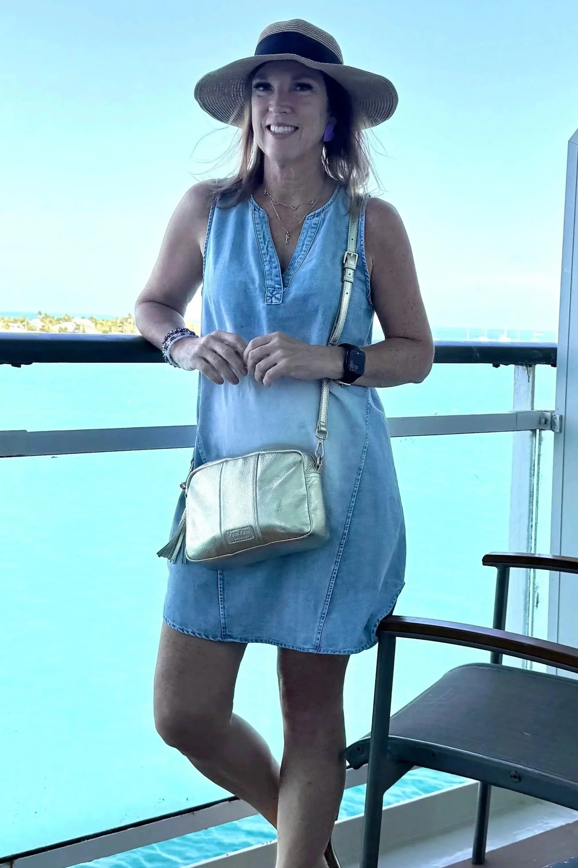 A woman wearing a wide-brimmed hat and denim dress standing on a balcony overlooking the ocean, with a blue sky in the background.