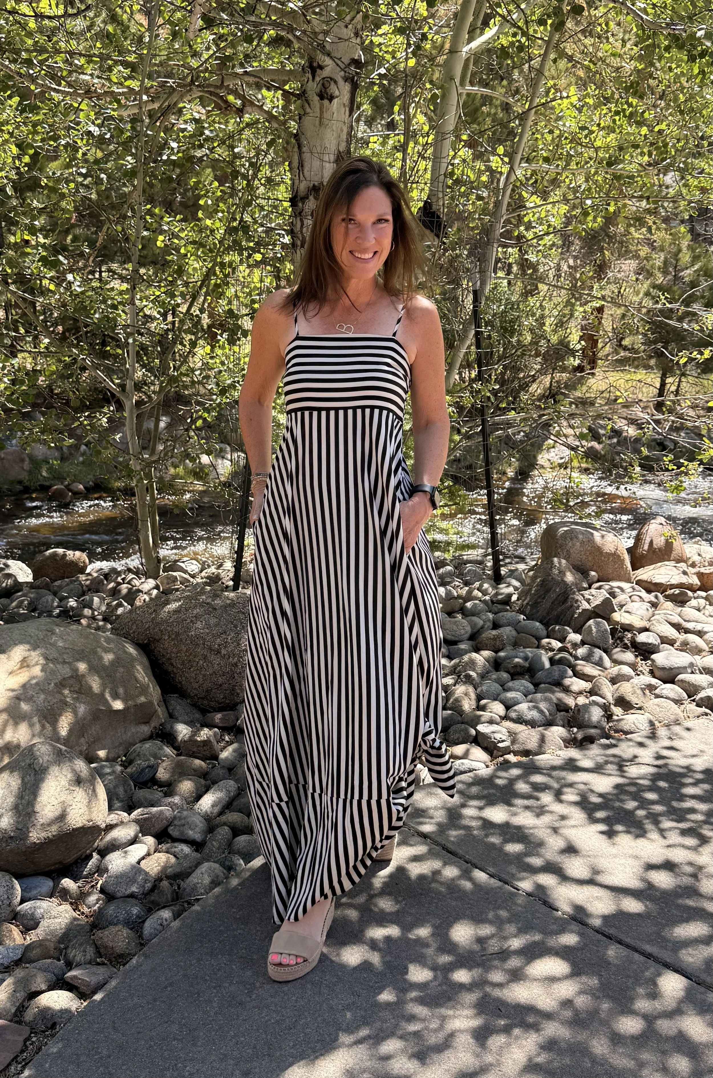 A woman with brown hair smiling outdoors in a wooded area, wearing a black and white striped sundress, standing on a path with rocks and trees in the background.