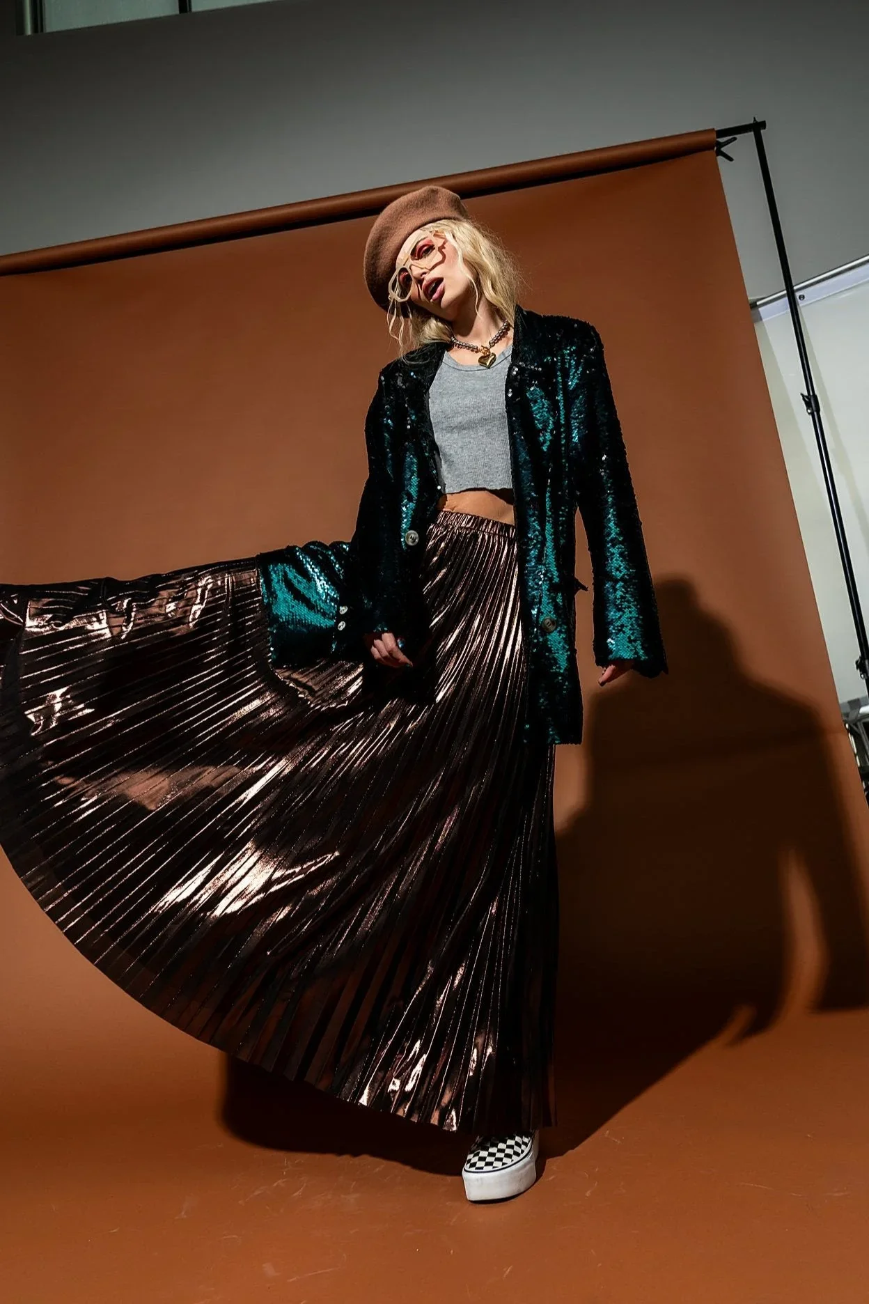 Person wearing a brown beret, glasses, a black shiny jacket, a gray crop top, a metallic pleated skirt, checkered platform shoes, posing in front of a brown backdrop.