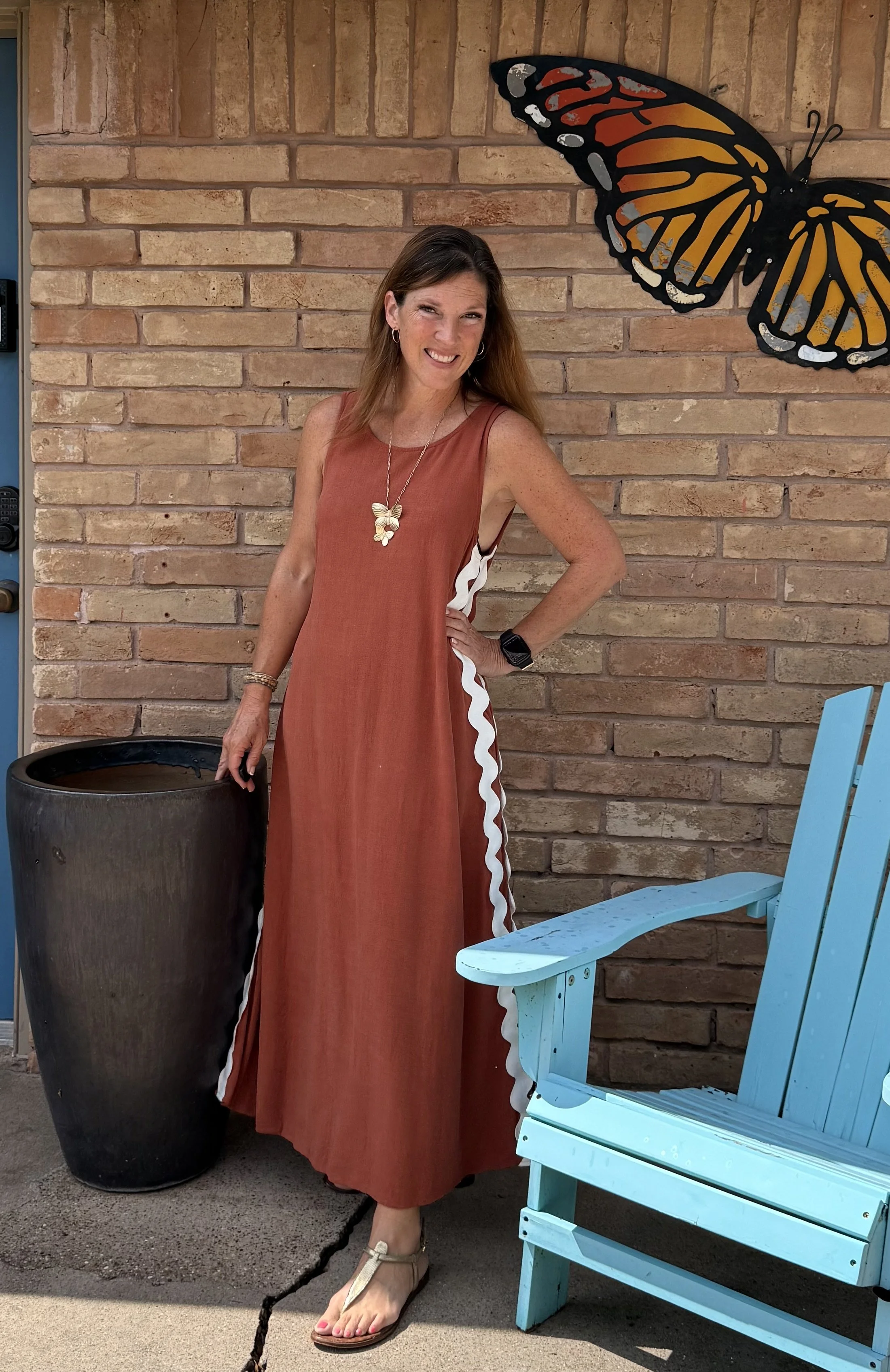 A woman in a long brown dress with white accents, sandals, and a butterfly necklace stands smiling beside a large black pot and a bright blue wooden chair, against a brick wall decorated with a large butterfly sculpture.