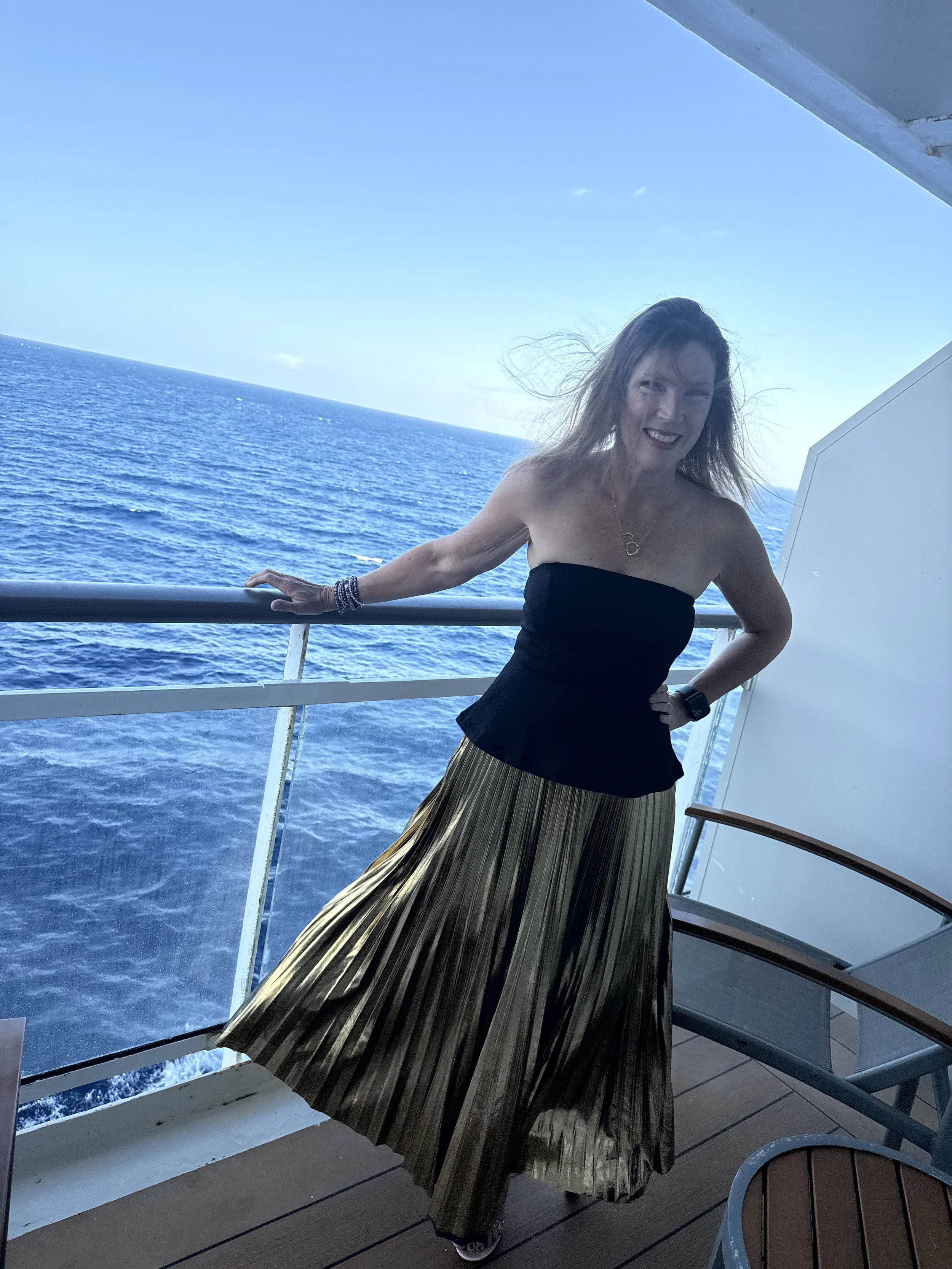 A woman with long hair wearing a black strapless top and a gold pleated skirt, smiling on a boat with ocean in the background.