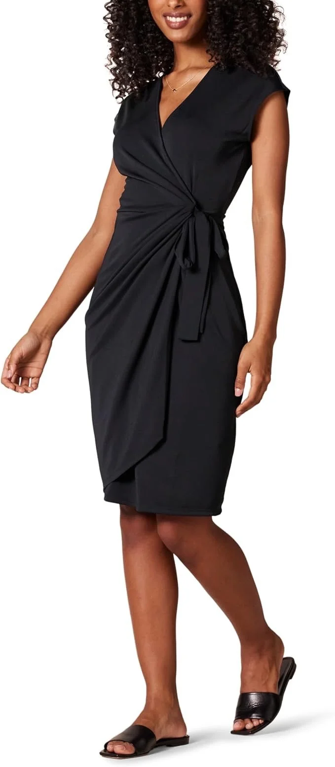 A woman in a black dress with a tied knot at the waist, wearing black open-toe sandals, smiling, against a white background.