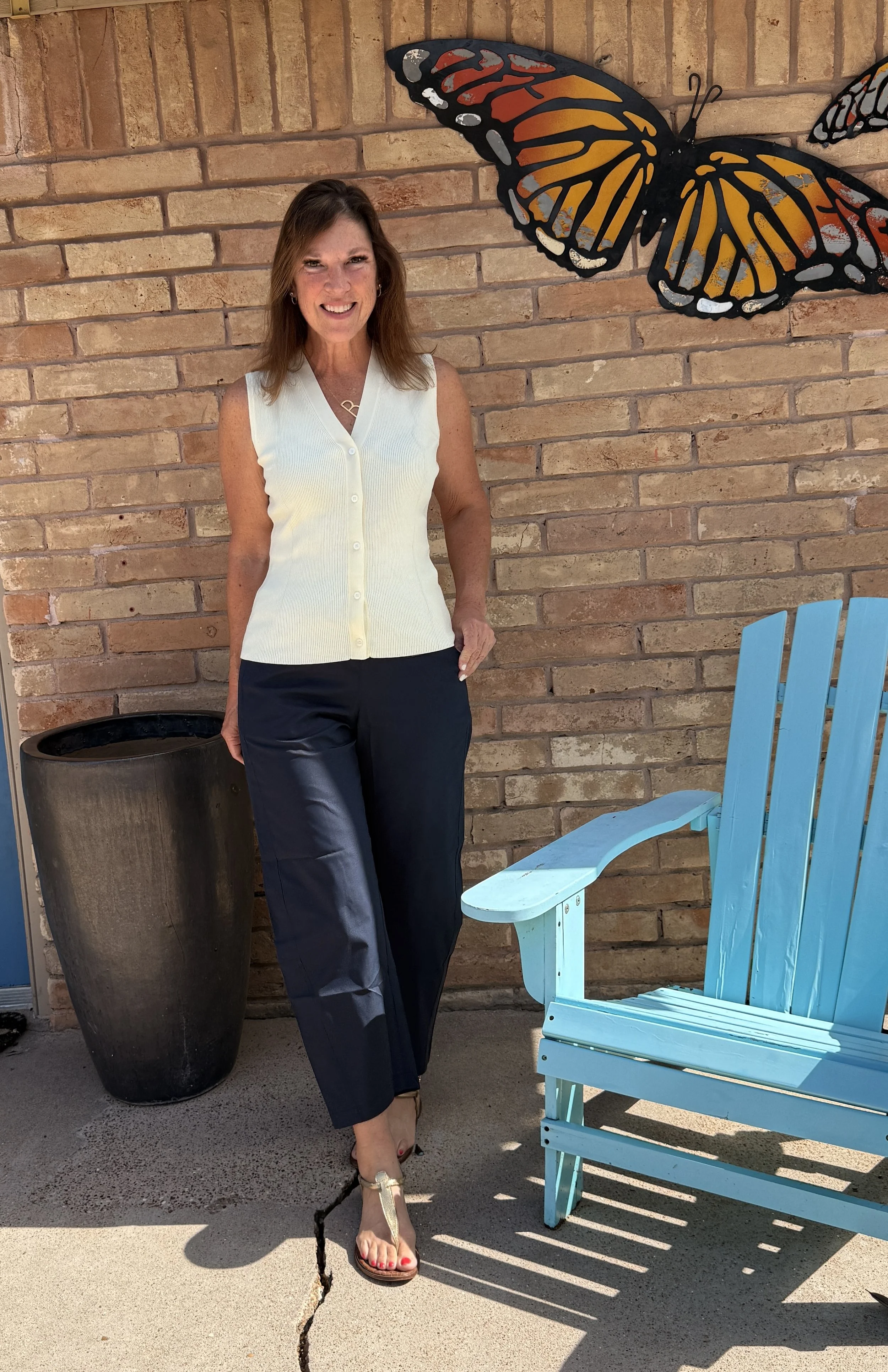 A woman standing next to a large black flowerpot, smiling and posing in front of a brick wall with metal butterfly art. She is wearing a sleeveless button-up top, black pants, sandals, and has shoulder-length brown hair.