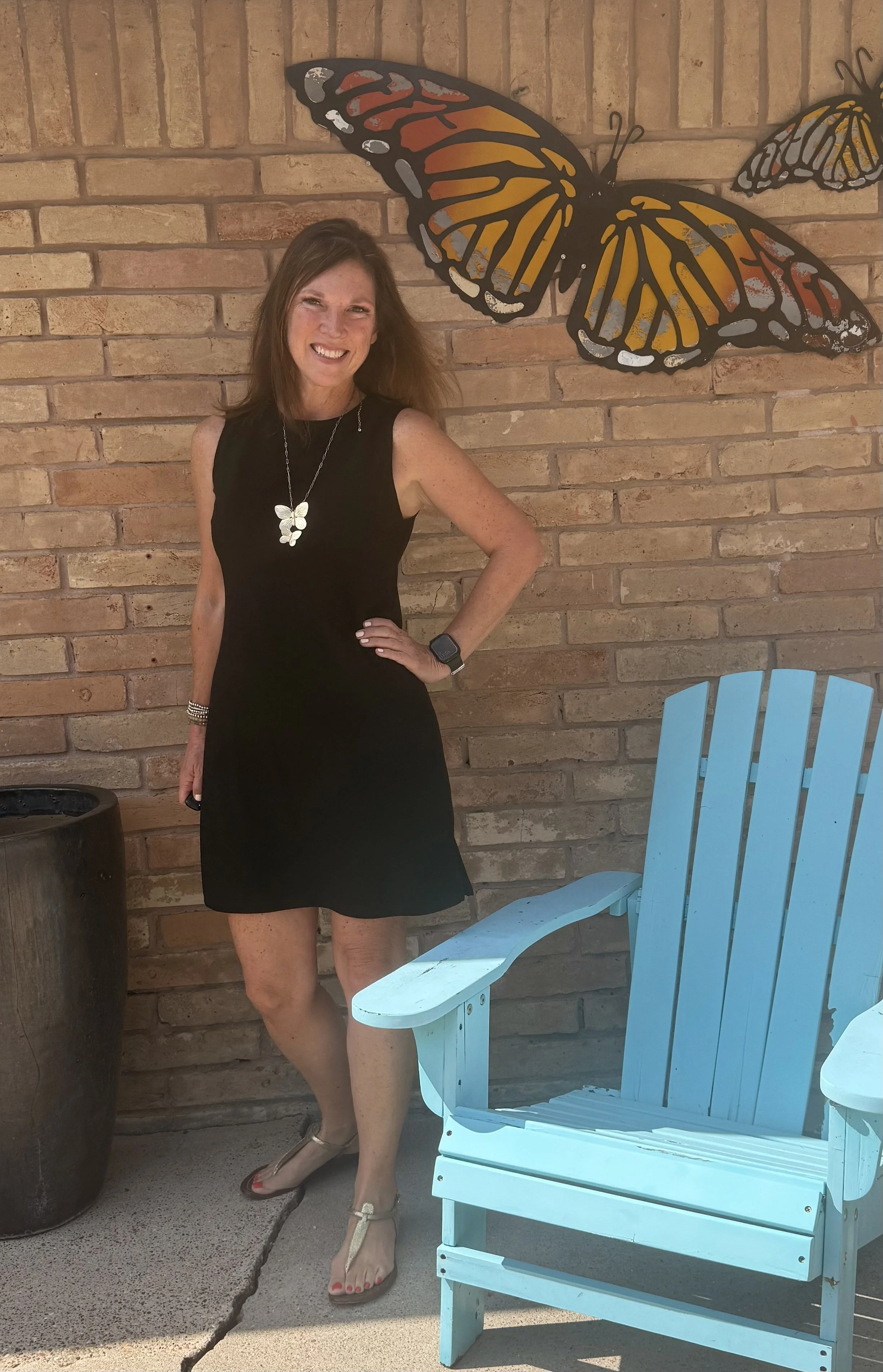 A woman with brown hair wearing a black sleeveless dress, butterfly necklace, and sandals posing with her hand on her hip beside a turquoise Adirondack chair against a brick wall with a large butterfly art piece.