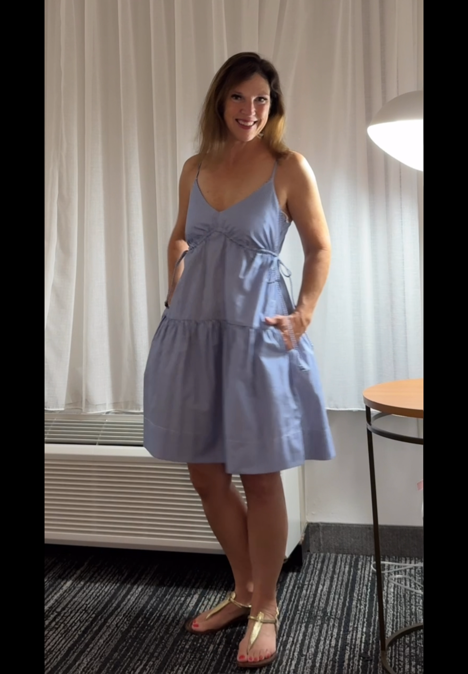 A woman is standing indoors in front of white curtains, wearing a light purple dress with spaghetti straps and yellow sandals. She has shoulder-length brown hair and is smiling at the camera.