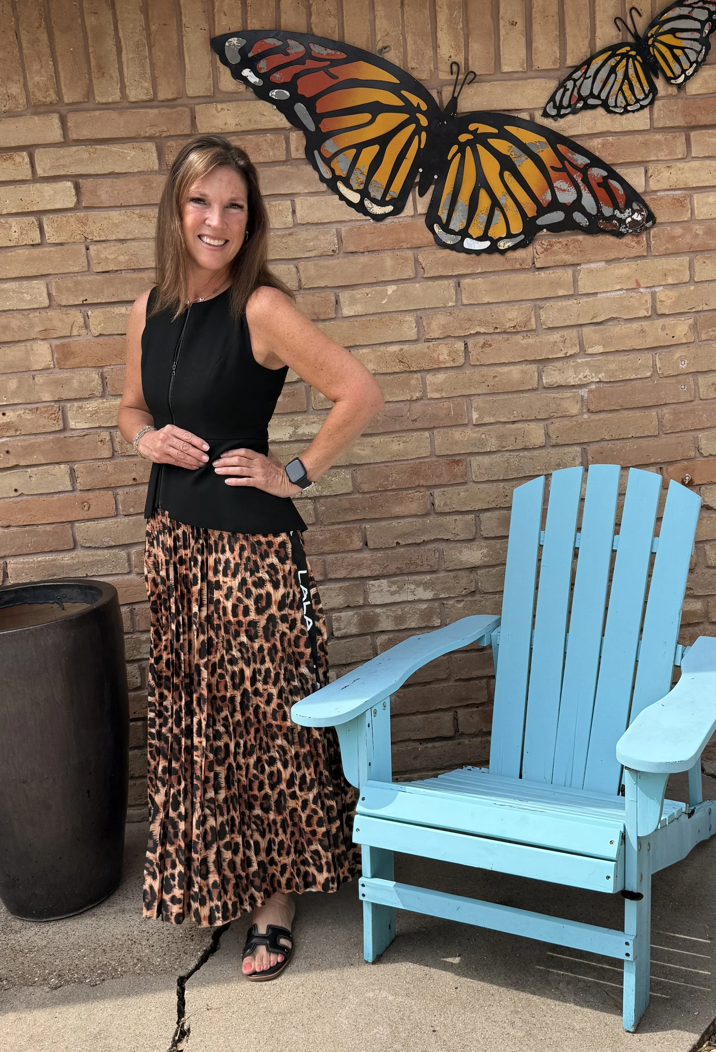 A smiling woman with brown hair, wearing a black sleeveless top, a leopard print long skirt, and sandals, posing with her right hand on her hip next to a bright blue wooden Adirondack chair, a tall black planter, and a brick wall decorated with two large butterfly wall art pieces.