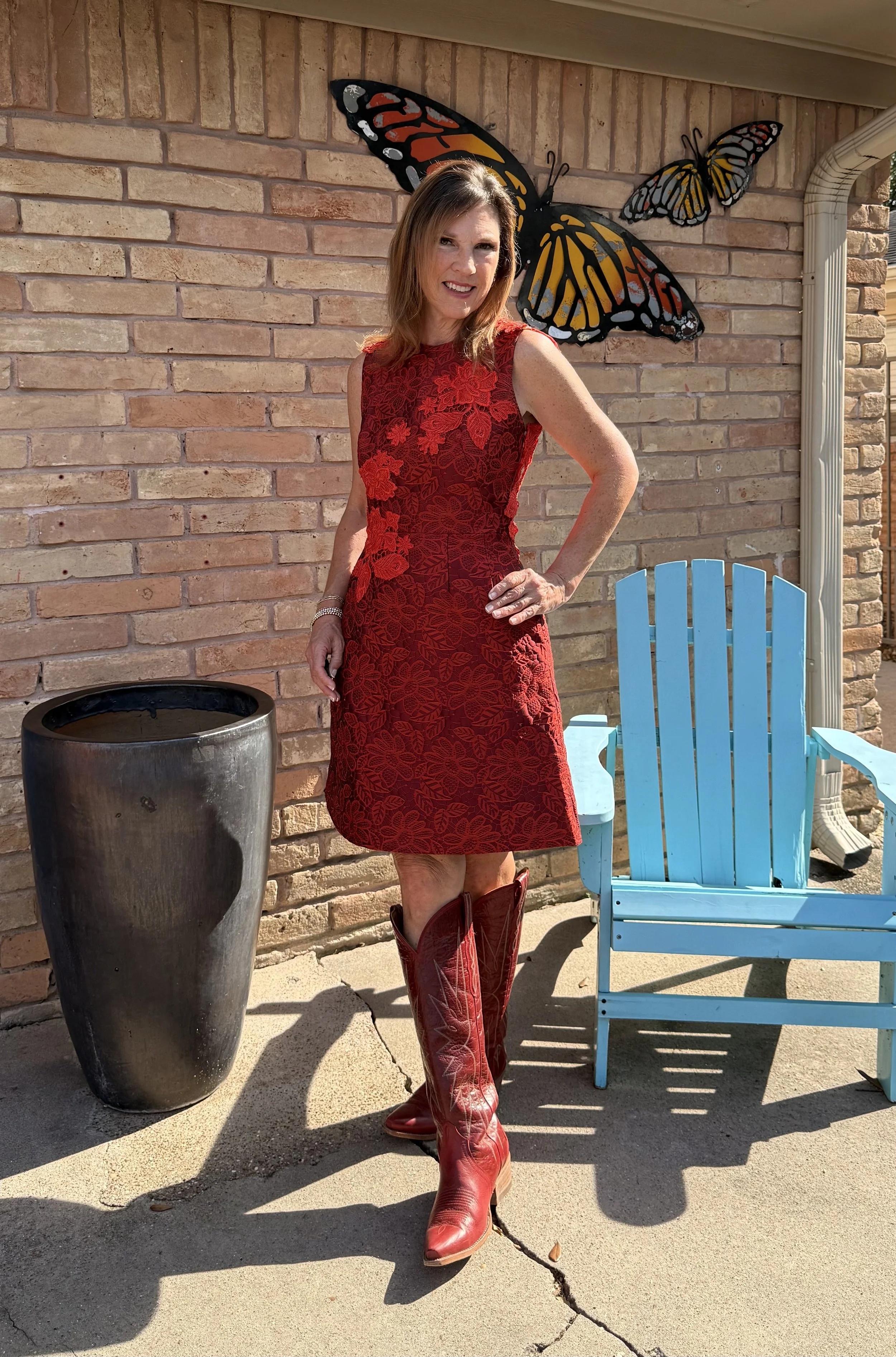 A woman in a red dress and cowboy boots standing outdoors in front of a brick wall with butterfly art decorations. She's smiling and has her hand on her hip, with a blue chair and large black planter nearby.