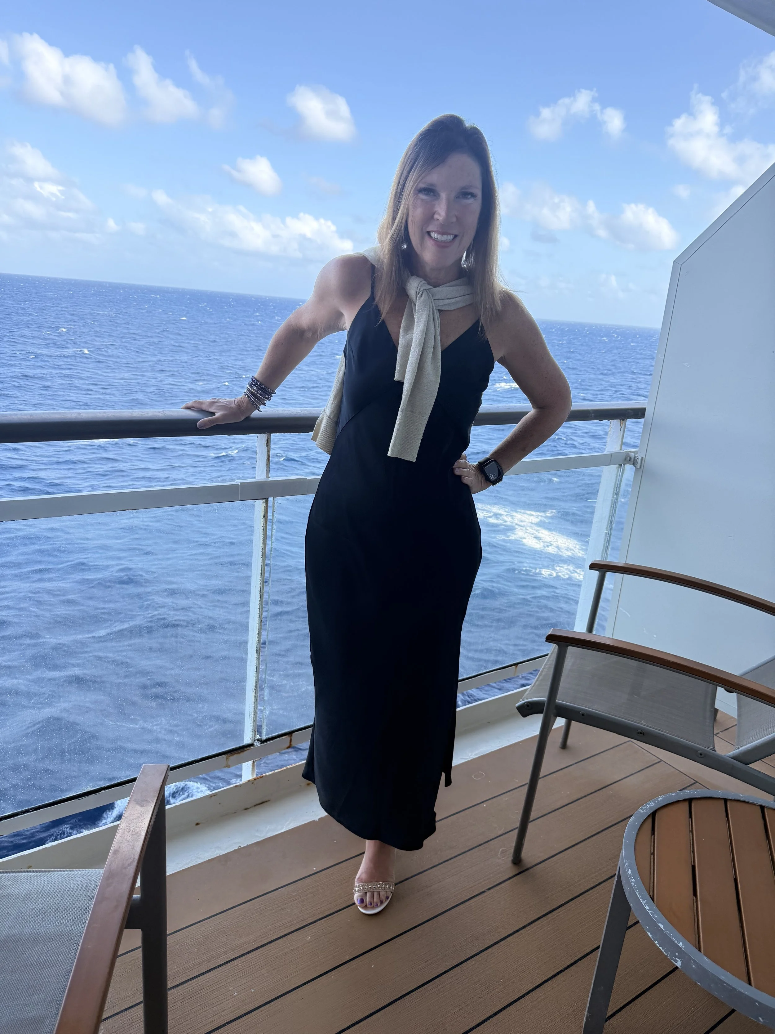 A woman in a black dress with a beige scarf draped around her neck, standing on the deck of a cruise ship with an ocean and cloudy sky in the background.