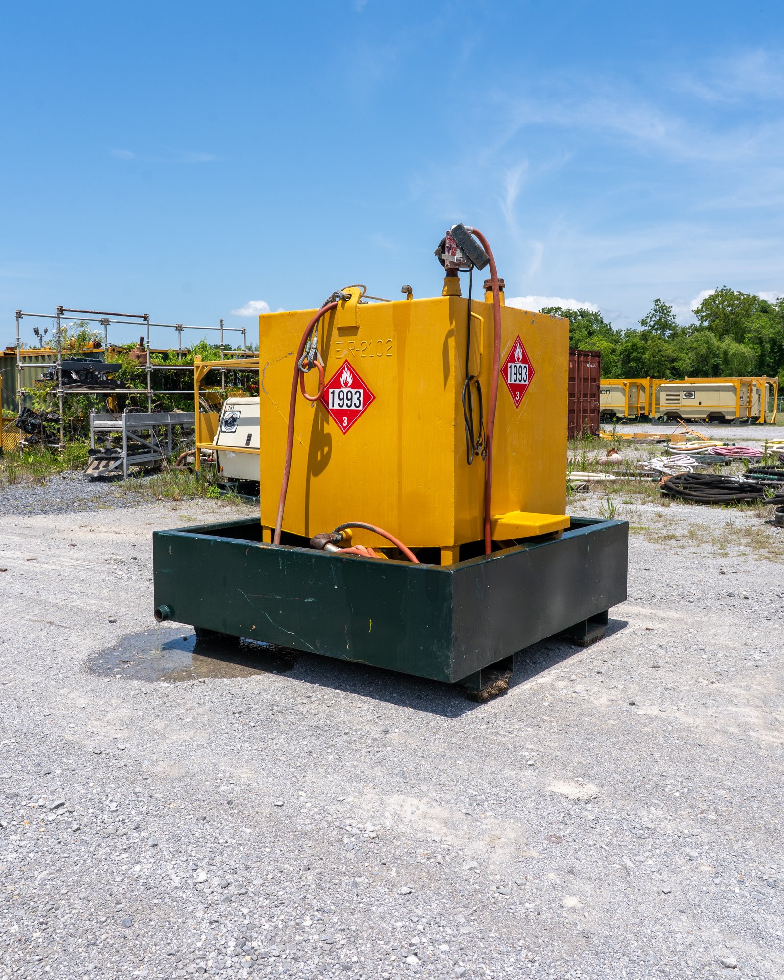 Large yellow metal container with hazard labels, connected to hoses, placed outdoors on gravel with trees and equipment in the background.