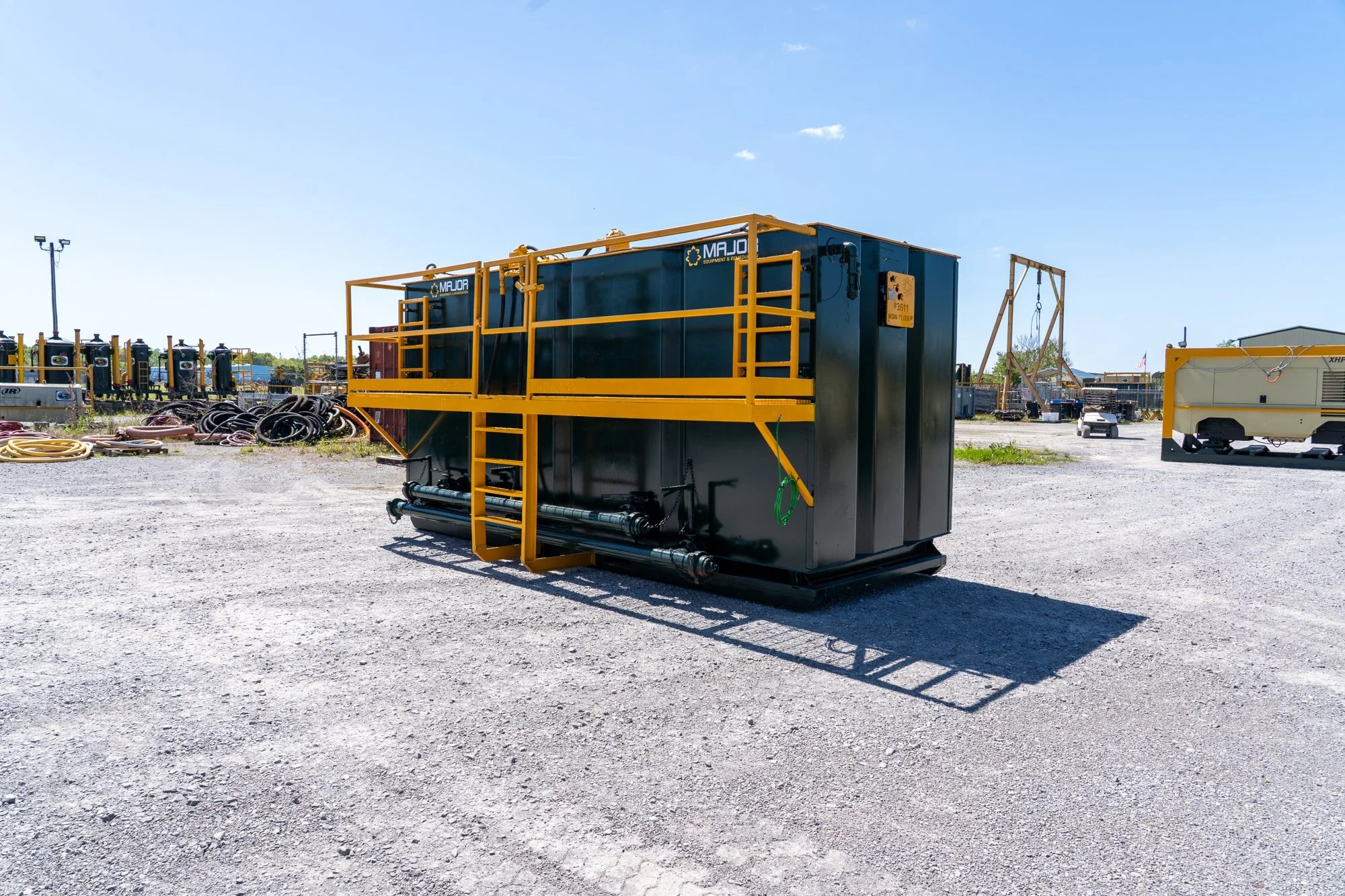 Large industrial black equipment container with yellow railing and stairs, situated outdoors on gravel ground, with other industrial equipment and hoses in the background under a clear blue sky.