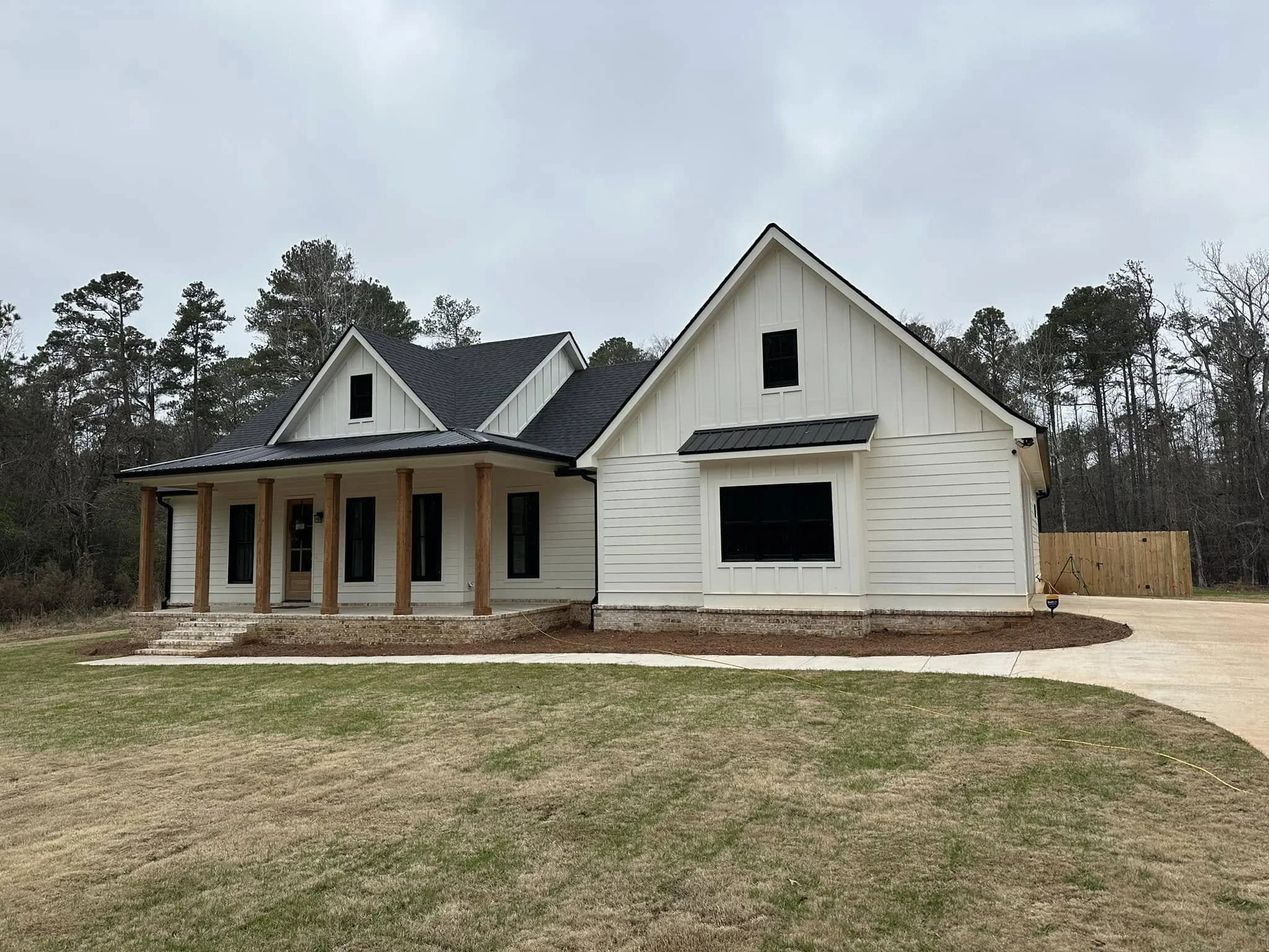 New white house with black roof, front porch with wooden columns, and a curved driveway, surrounded by a grassy lawn and trees in the background.