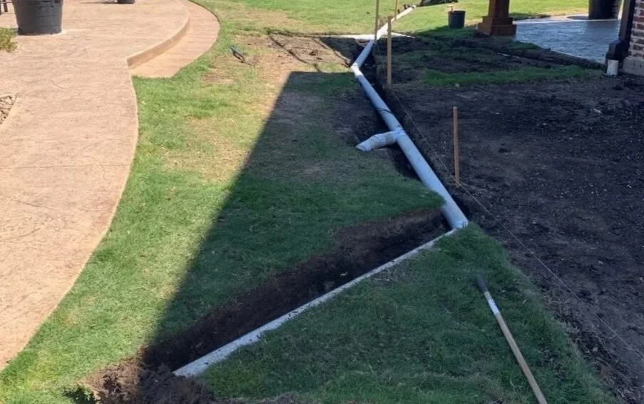 A partially completed landscaping project with a trench along the edge of a lawn, containing white PVC pipes and some wires, possibly for drainage or irrigation, next to a house.