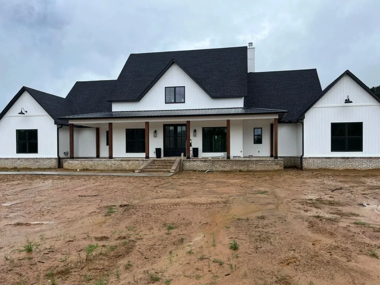 A modern house with a black roof, white exterior, and a front porch with wooden columns, set on a dirt yard.