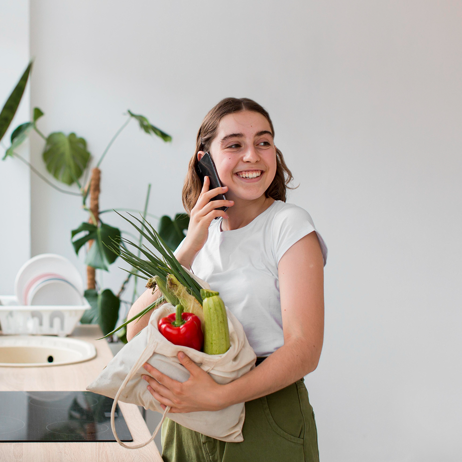A woman smiling while talking on the phone and holding a grocery bag with vegetables including a red bell pepper, zucchini, and green onion in a bright kitchen.