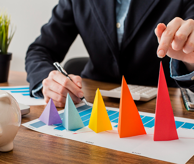Person in business attire pointing at colorful paper cones on a chart on a wooden desk.