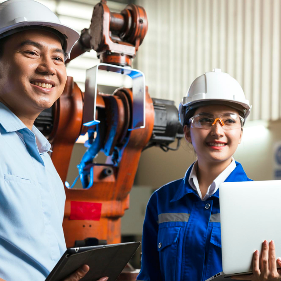Two engineers, a man and a woman, in safety helmets and glasses, working with a robotic arm in an industrial setting.