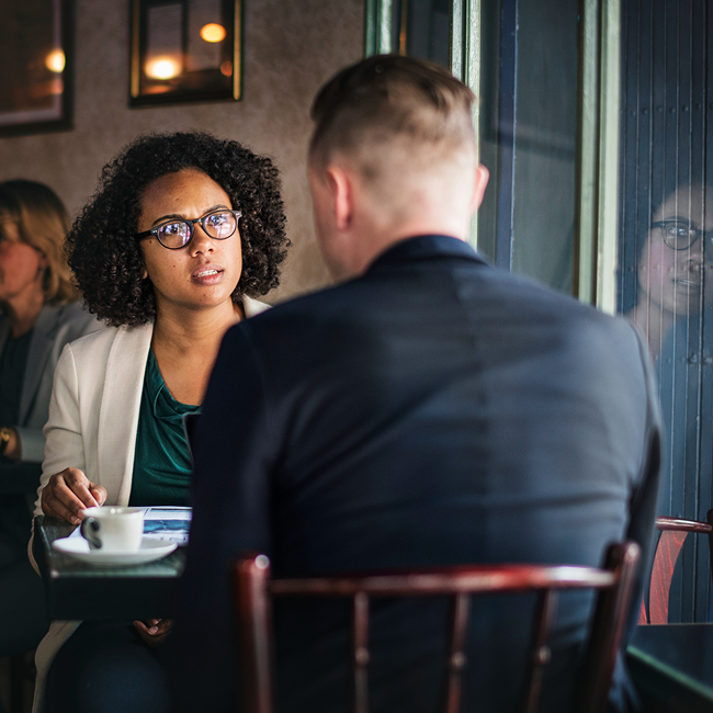 Two people having a serious conversation at a restaurant table, with a woman wearing glasses and a white blazer looking at a man in a dark suit. Other patrons are visible in the background.