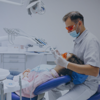 Dentist wearing red protective glasses and gloves examining a patient in a dental chair.