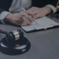 A judge's gavel resting on a sound block on a desk, with a person writing on a notepad in the background.