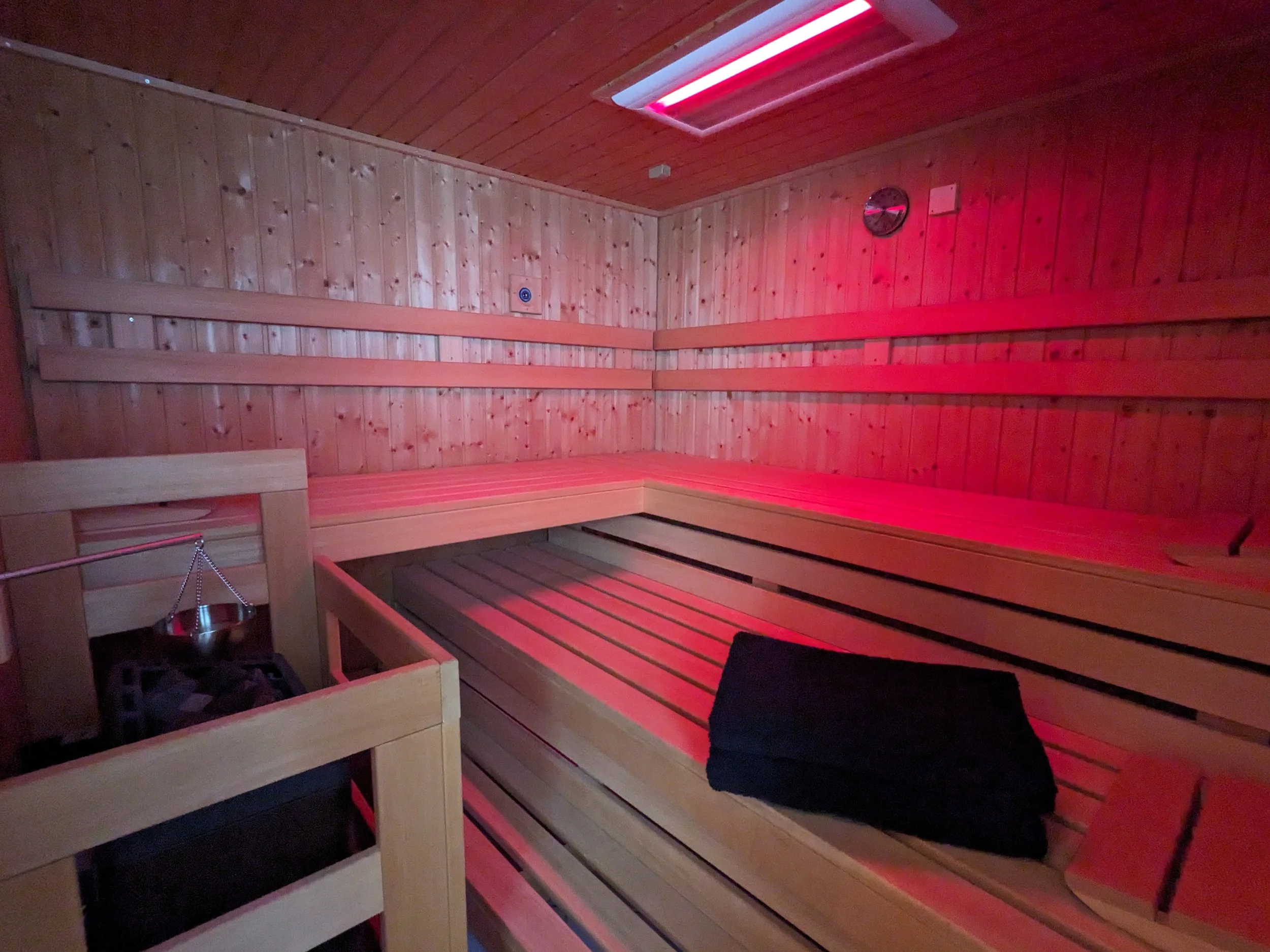Innenraum einer Sauna mit Holz-Wänden und -Bänken, rotes Licht. Es befindet sich eine schwarzes Kissen auf einer der Bänke, eine Glastür und eine Uhr an der Wand.