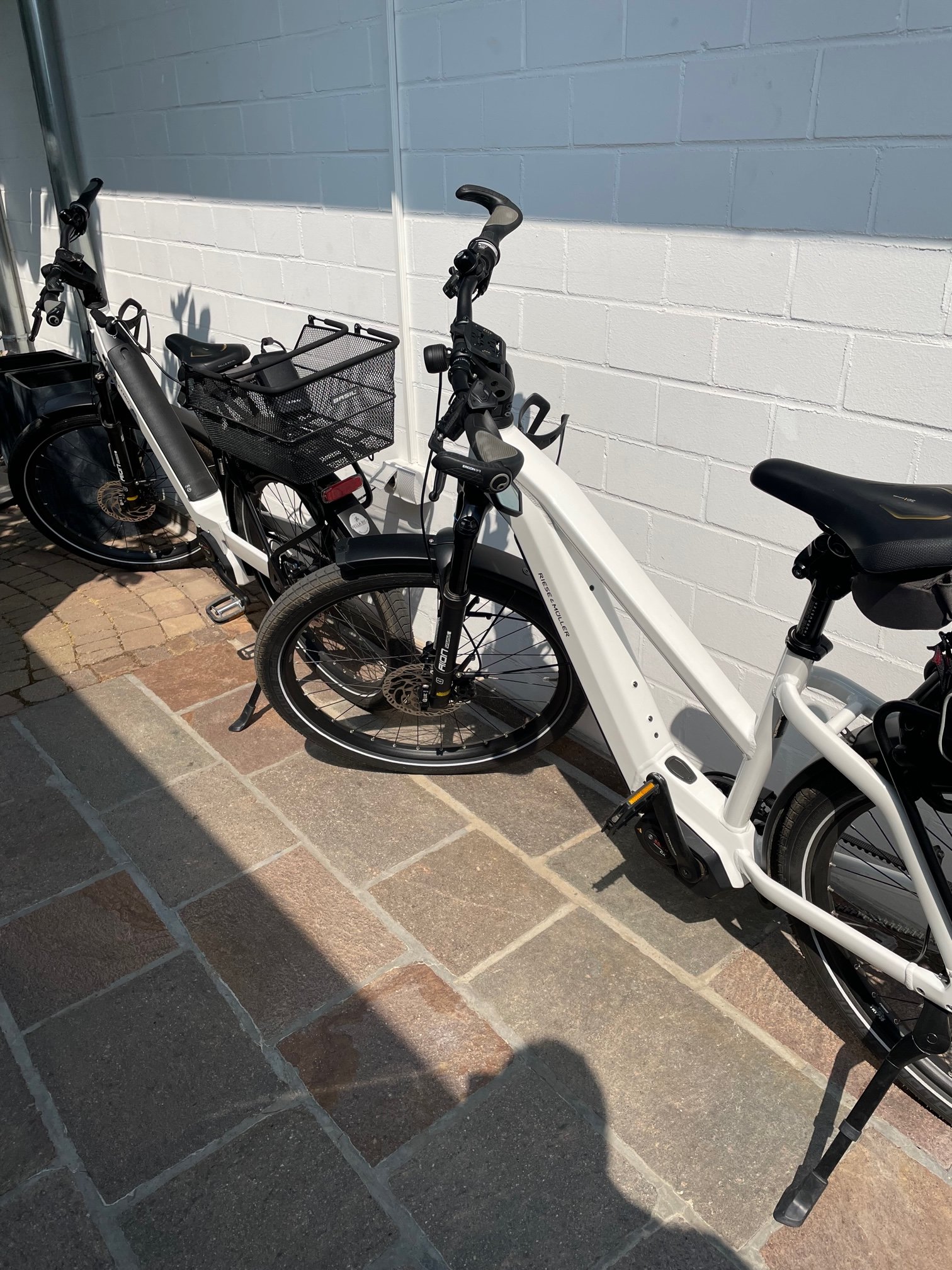 Blick auf zwei elektrische Fahrräder, eines mit hinterer Tasche, die vor einer weißen Wand mit grauen Ziegeln abgestellt sind.