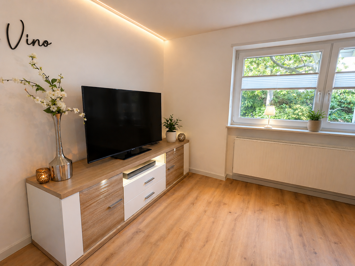 Modernes Wohnzimmer mit Fernseher auf Holz- und Weißer Kommode, Vase mit weißen Blumen, kleinen Pflanzen, Fenster mit Blick auf grüne Bäume und indirekte Deckenbeleuchtung.