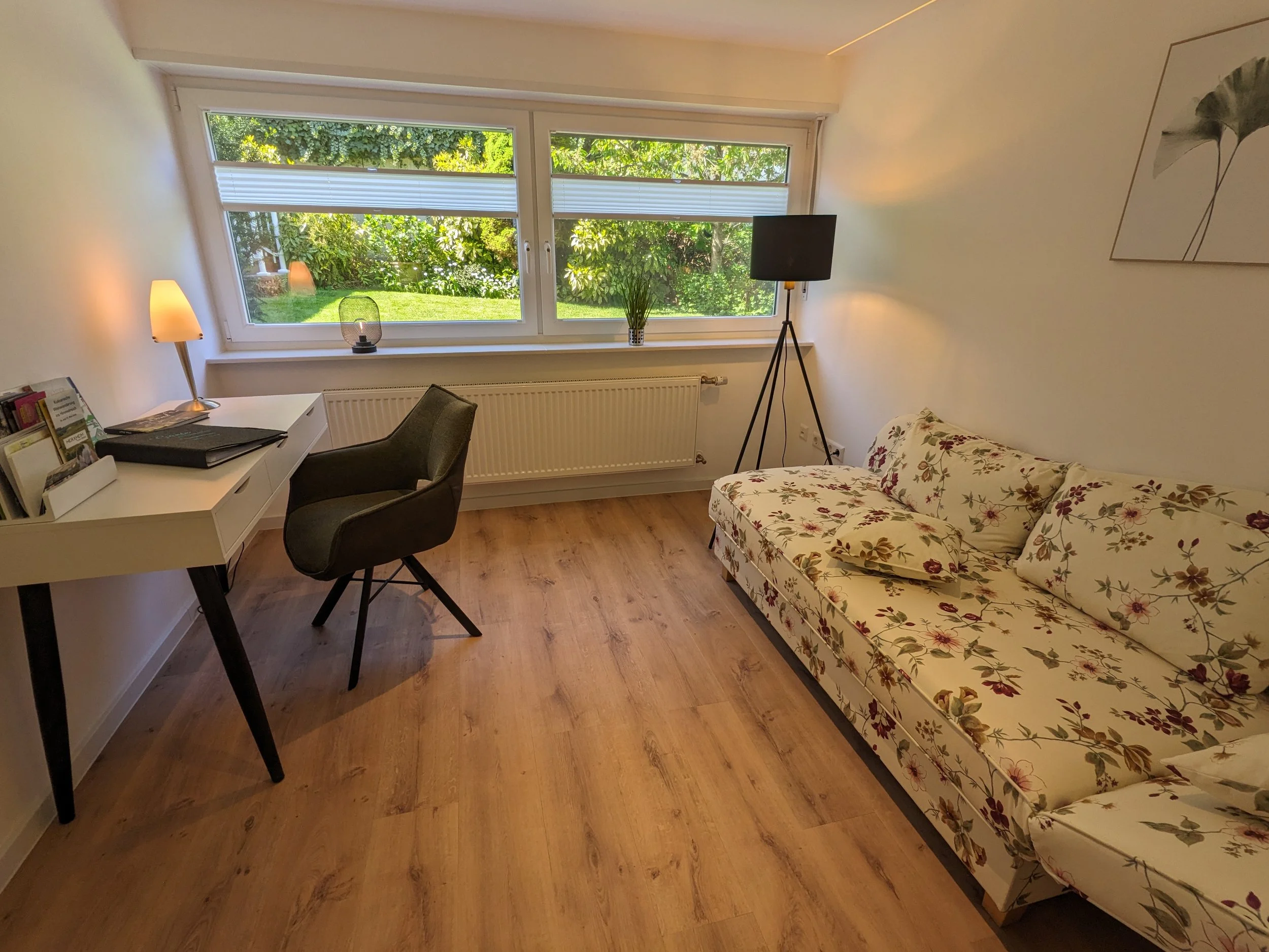 Ein Zimmer mit einem Schreibtisch, einem Stuhl, einem Sofa mit Blumendruck, einem Fernseher, einer Lampe, einem Bücherregal und einer großen Fensterfront mit Blick ins Grüne.