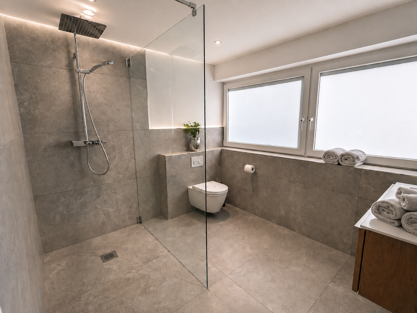 Modernes Badezimmer mit begehbarer Dusche, Wand-WC, großen Fenstern, grauen Fliesen, weißen Handtüchern und einer Pflanze auf der Ablage.