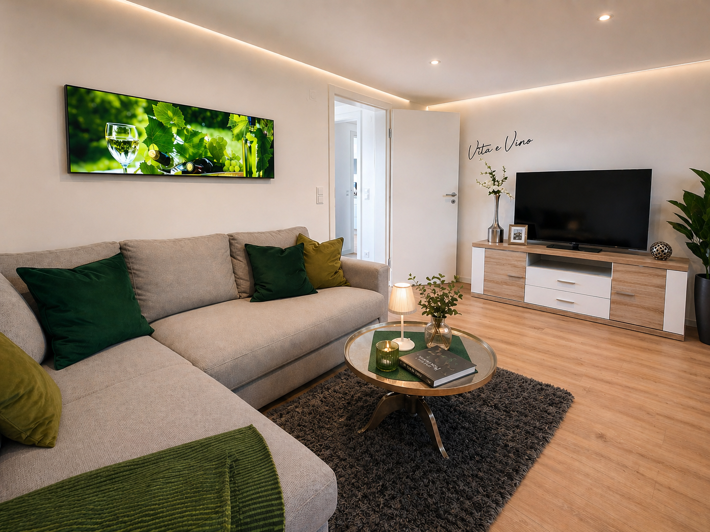 Modernes Wohnzimmer mit grauer Couch, grünen Kissen, einem runden Couchtisch mit einer kleinen Lampe, Pflanzen und einem großen Fernseher auf einem Holz-Tisch. Es gibt einen offenen Tür in den nächsten Raum und Wanddekoration mit der Aufschrift 'Vita e Vino'.