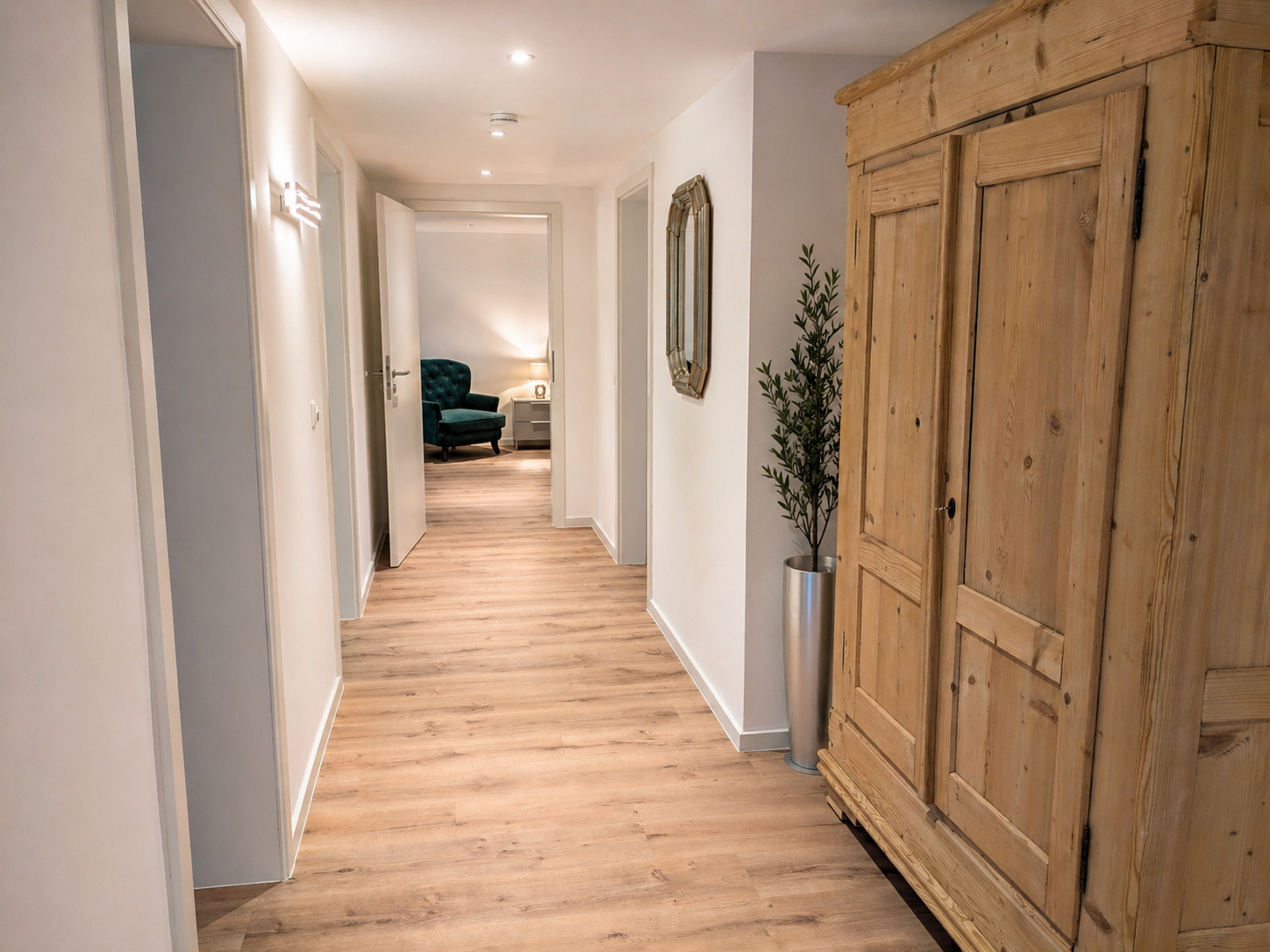 Heller Flur mit Holzboden, weißen Wänden, einem großen Holzschrank, einem Spiegel an der Wand, einer Topfpflanze in einem silbernen Topf, einem offenen Türrahmen und einem gemütlichen Wohnzimmer im Hintergrund mit einem grünen Sessel und einem Nachtt